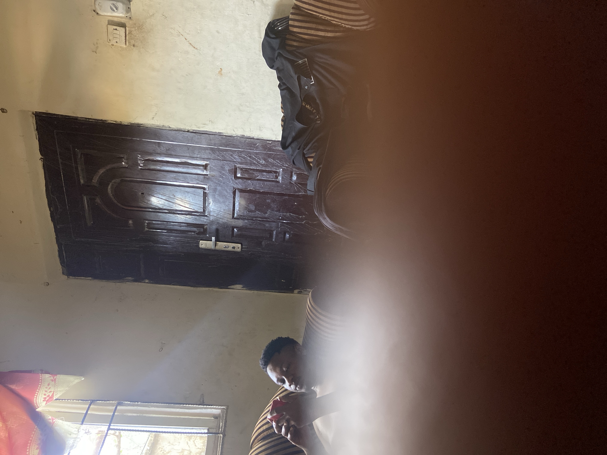 The image, taken indoors and likely in a private residence in Mararaba, Nigeria, shows a partially obstructed view of a room. The room appears sparsely furnished. An interior door of dark, possibly varnished wood, is visible. Above the door, the wall is painted a light, neutral color with an electrical outlet. The lower part of the frame is mostly obscured by a hand and the lower part of a human. What may be a window is seen in the bottom left corner. The light outside the window suggests it's daytime. The image's overall quality is poor due to the obstruction and possibly blurred focus.