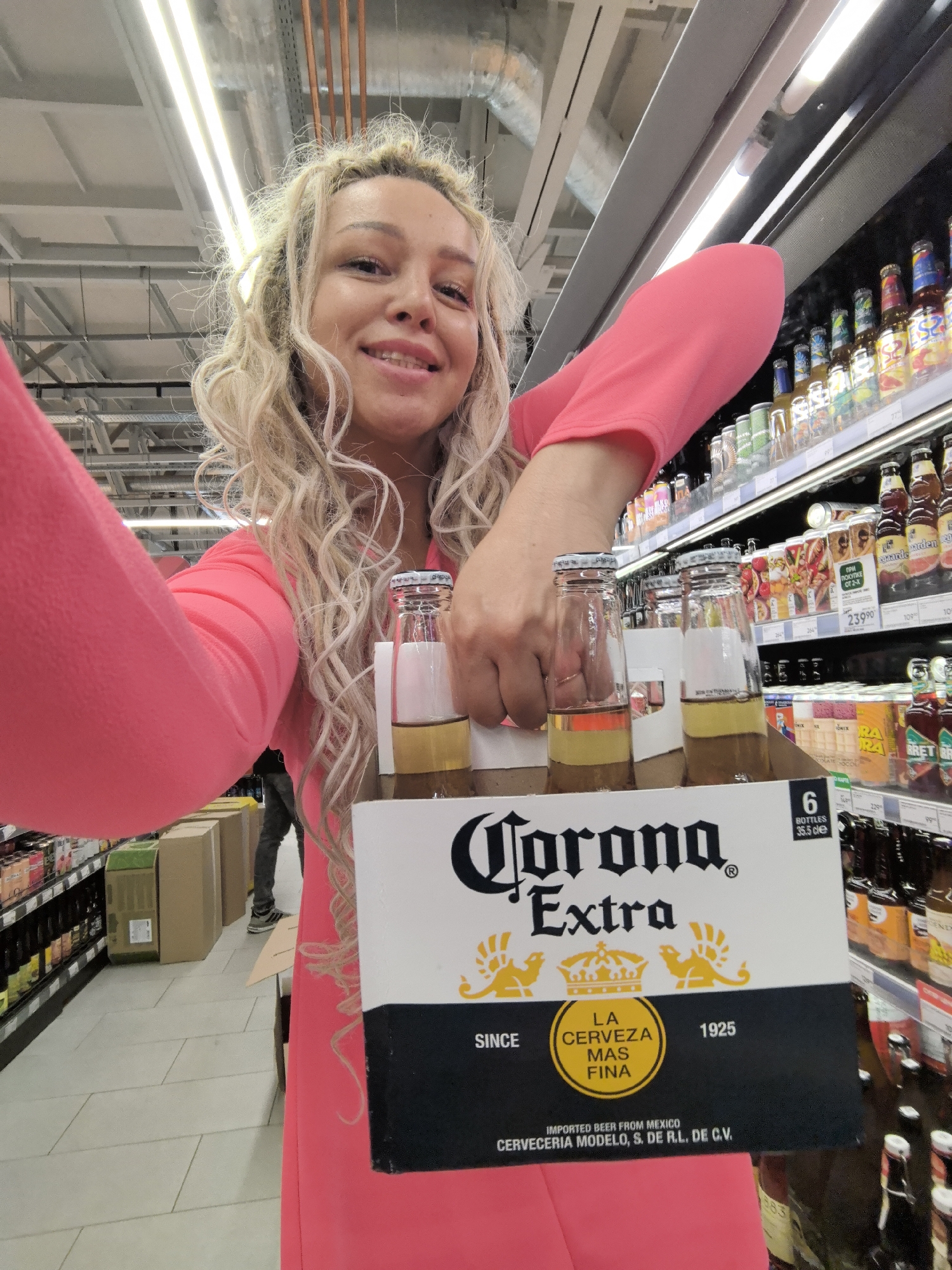 A woman with blonde curly hair is holding a six-pack of Corona Extra beer in a store. She is wearing a bright pink dress. The beer bottles are visible in the foreground, and the woman's face is smiling at the camera. In the background, there are shelves stocked with various beverages, indicating the location is a supermarket or a liquor store. There are also some cardboard boxes stacked in the aisle behind her. The lighting suggests the interior of a well-lit retail establishment. The image does not contain information to determine the location in Nizhny Novgorod, Russia.