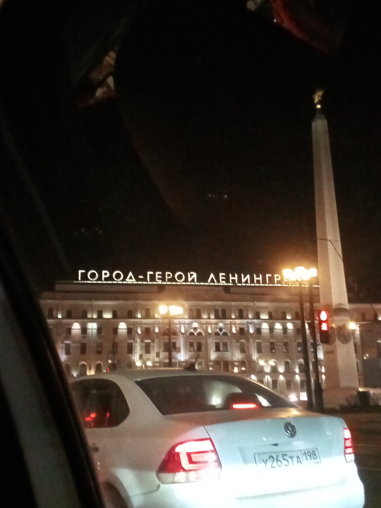 The image depicts a nighttime urban scene in Saint Petersburg, Russia, viewed from a low angle, likely from inside a vehicle due to the dark framing elements on the left and top. In the foreground, the rear of a white sedan, displaying a Russian license plate "У 265ТА 198" (indicating the Saint Petersburg region), is visible with its brake lights illuminated.
To the right of the sedan stands a tall, light-colored obelisk monument, topped with a golden sculptural element. A traffic light pole with a red signal is positioned to the left of the obelisk.
In the background, a large, multi-story building with illuminated windows is visible. Across its rooftop, large, white, illuminated Cyrillic letters spell out "ГОРОД-ГЕРОЙ ЛЕНИНГРАД" (HERO CITY LENINGRAD). This building is identified as the Moscow Railway Station, and the monument is the Obelisk "Hero City Leningrad", both located on Vosstaniya Square in Saint Petersburg.
The environment is illuminated by artificial streetlights and building facade lighting. No individuals are discernible within the frame.