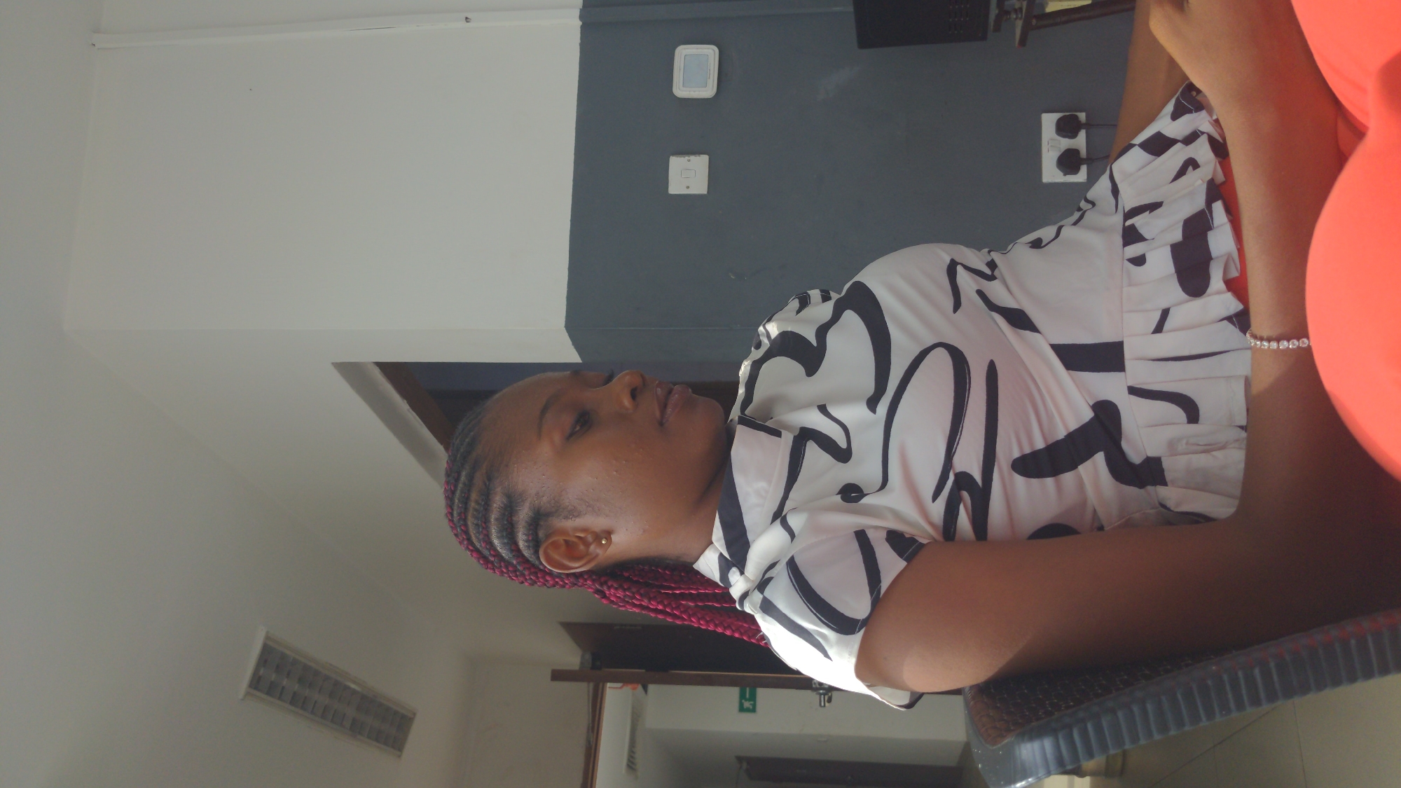 An indoor, medium shot captures a woman seated, looking slightly to her left, with the image rotated 90 degrees counter-clockwise. The woman, with brown skin, wears her hair in tight red/maroon braided cornrows. She has a subtle gold stud earring in her right ear. Her expression is calm and neutral.
She is dressed in a white shirt adorned with bold, abstract black patterns. Her arms are bent, resting on a dark, possibly woven or ribbed surface, which appears to be a table or armrest. On her right wrist, she wears a delicate bracelet with small, white, possibly beaded or jeweled accents. A bright red or orange object, perhaps a cushion or another person's clothing, is partially visible to her right.
The background features a two-toned wall; the upper section is white, and the lower section is dark grey. Several electrical fixtures are mounted on the dark grey part of the wall: two white electrical outlets with black plugs inserted, and above them, a white square object, possibly a thermostat or light switch. Further left on the white wall, an air vent is visible. A partially obscured green emergency exit sign (showing the 'E' symbol) is visible on a lower wall section to her left. The overall setting appears to be a modern interior in Abuja, Nigeria, possibly a cafe, office, or residential space. The lighting suggests it is daytime.