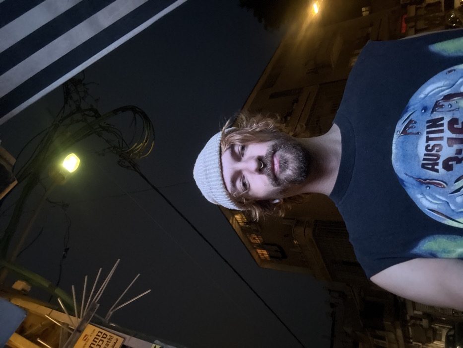 A man with a beard and a gray beanie looks directly at the camera, facing slightly downward. He wears a black t-shirt with "AUSTIN 3:16" visible on a circular design. The background features a dimly lit street with a street lamp and building facades, mostly dark. There are also wires and a partially visible awning or structure with black and white stripes in the upper left. The scene appears to be at night.