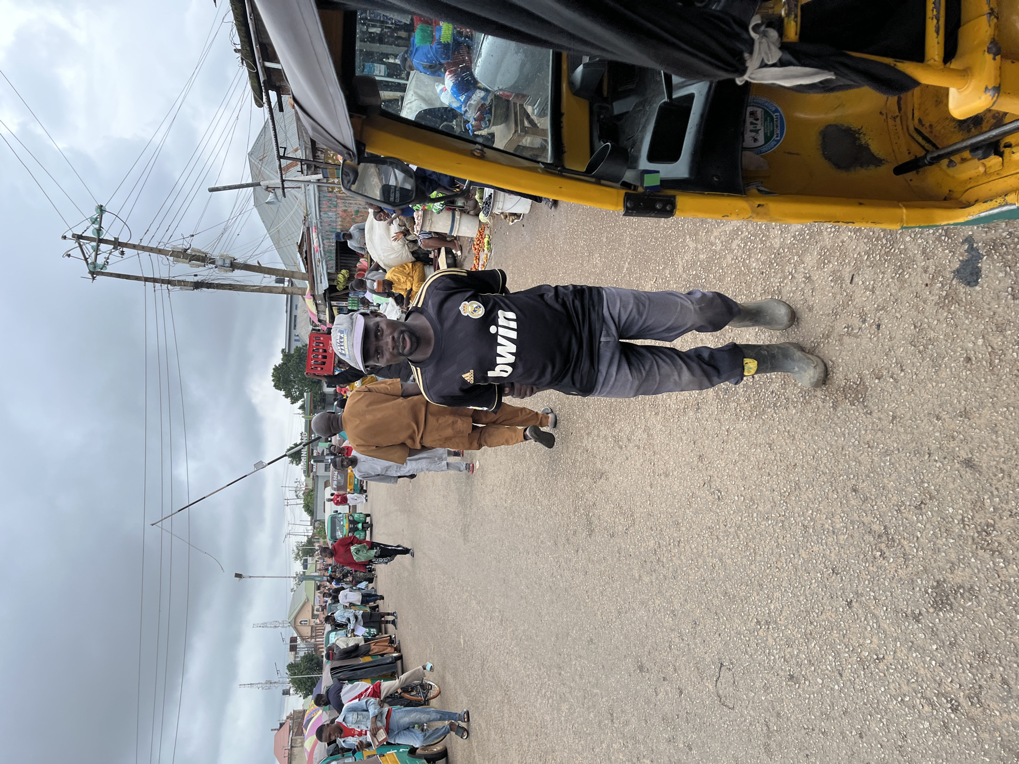 In Gura Top, Nigeria, a man wearing a black Adidas t-shirt with "bwin" written on it and gray pants stands in the foreground, facing the camera. He wears dark rubber boots. To his right, the yellow passenger compartment of a three-wheeled auto-rickshaw is visible, with a black cover obscuring part of the interior. The background depicts a busy street scene with numerous people, auto-rickshaws, market stalls, and overhead power lines. The sky is overcast.