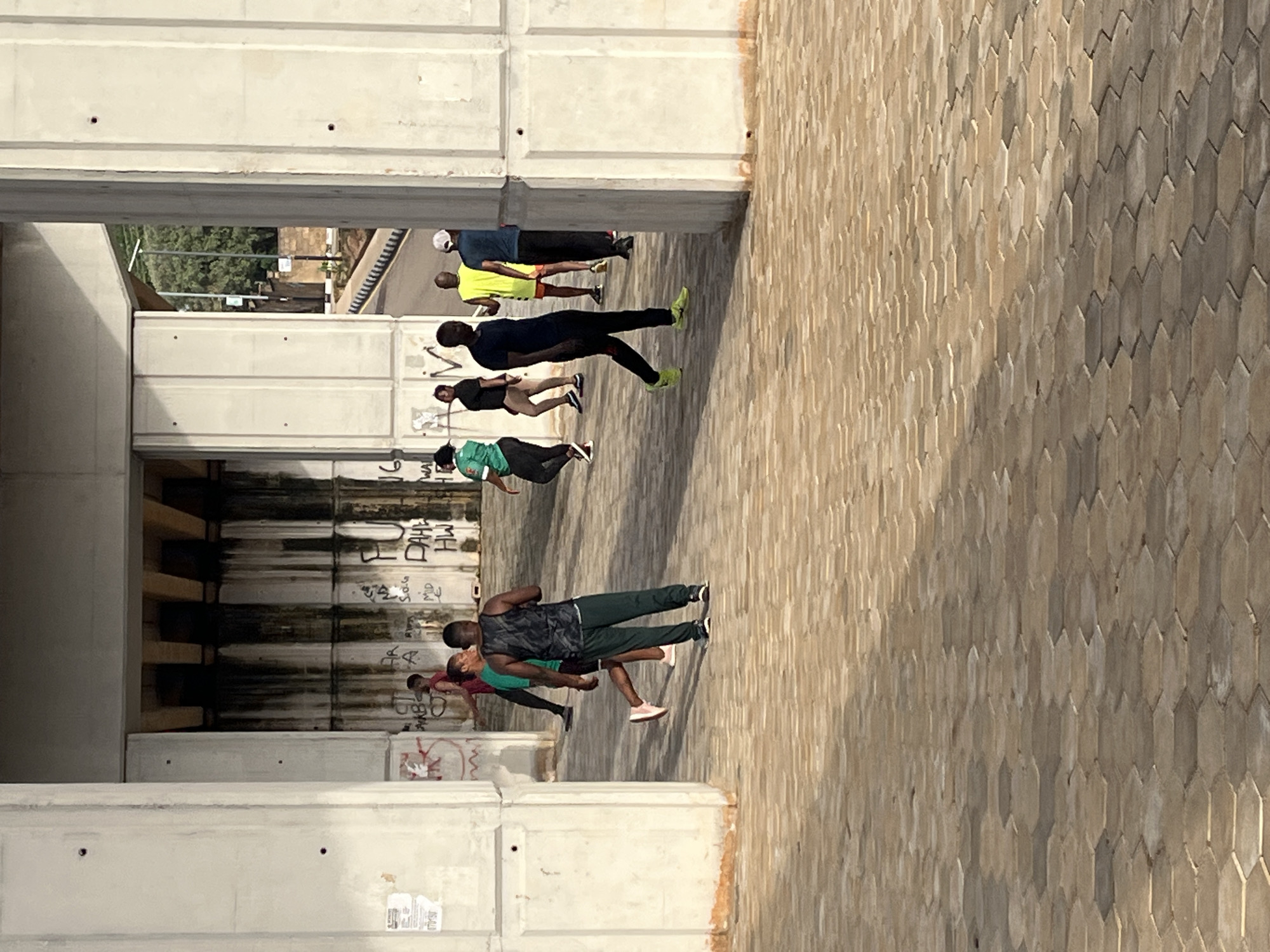 A group of people are exercising under a concrete overpass in Jos, Nigeria. The overpass pillars are made of concrete and show signs of graffiti. The ground is paved with hexagonal tiles. In the background, a road curves through a green, vegetated area. The people are dressed in athletic wear, suggesting they are engaged in a fitness activity. One person in the foreground is wearing a black tank top and dark pants, with bright yellow shoes. Another person is wearing a green shirt and dark pants. Several other individuals are visible further back, some running or walking. The lighting suggests it is daytime, possibly early morning or late afternoon, with the sun casting shadows.