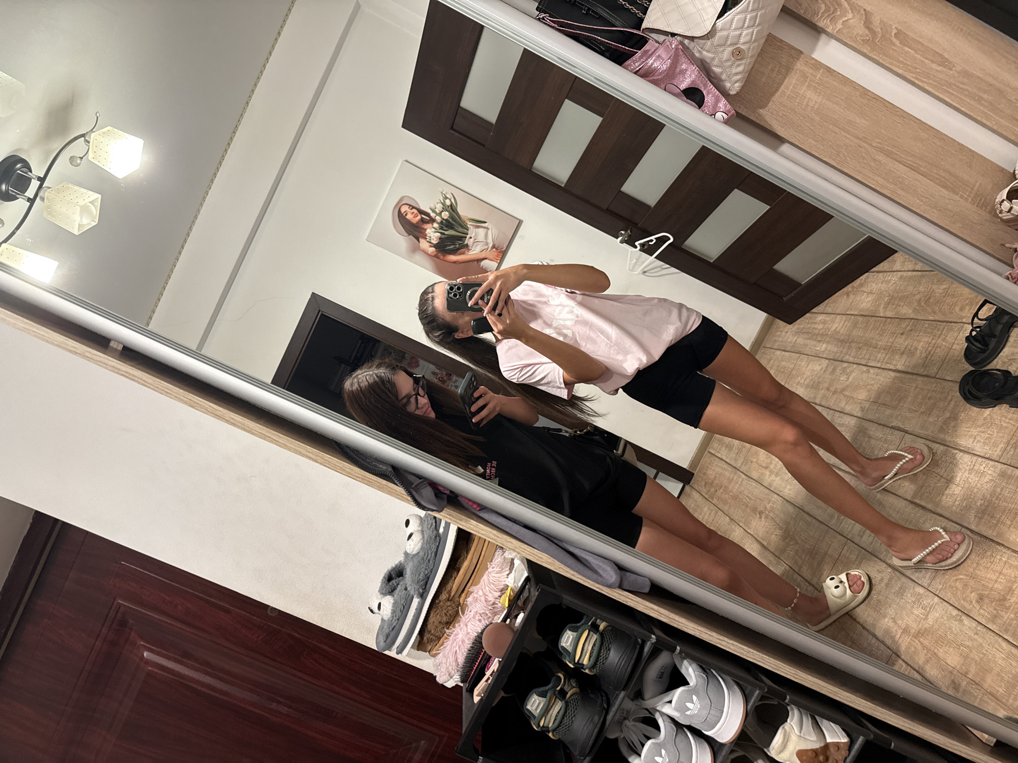 Two young women are taking a mirror selfie in a room. The woman on the left is wearing glasses, a black t-shirt, and black shorts. She is looking at her phone. The woman on the right is wearing a pink t-shirt and black shorts and is also looking at her phone. They are both standing on a wooden floor. To the left of the women is a shoe rack with several pairs of shoes and slippers. Behind the women, there is a wooden door and a painting on the wall. To the right of the women, there are more doors and some bags hanging on hooks. The lighting in the room is bright, and it appears to be daytime. The image was taken in Chișinău, Moldova.