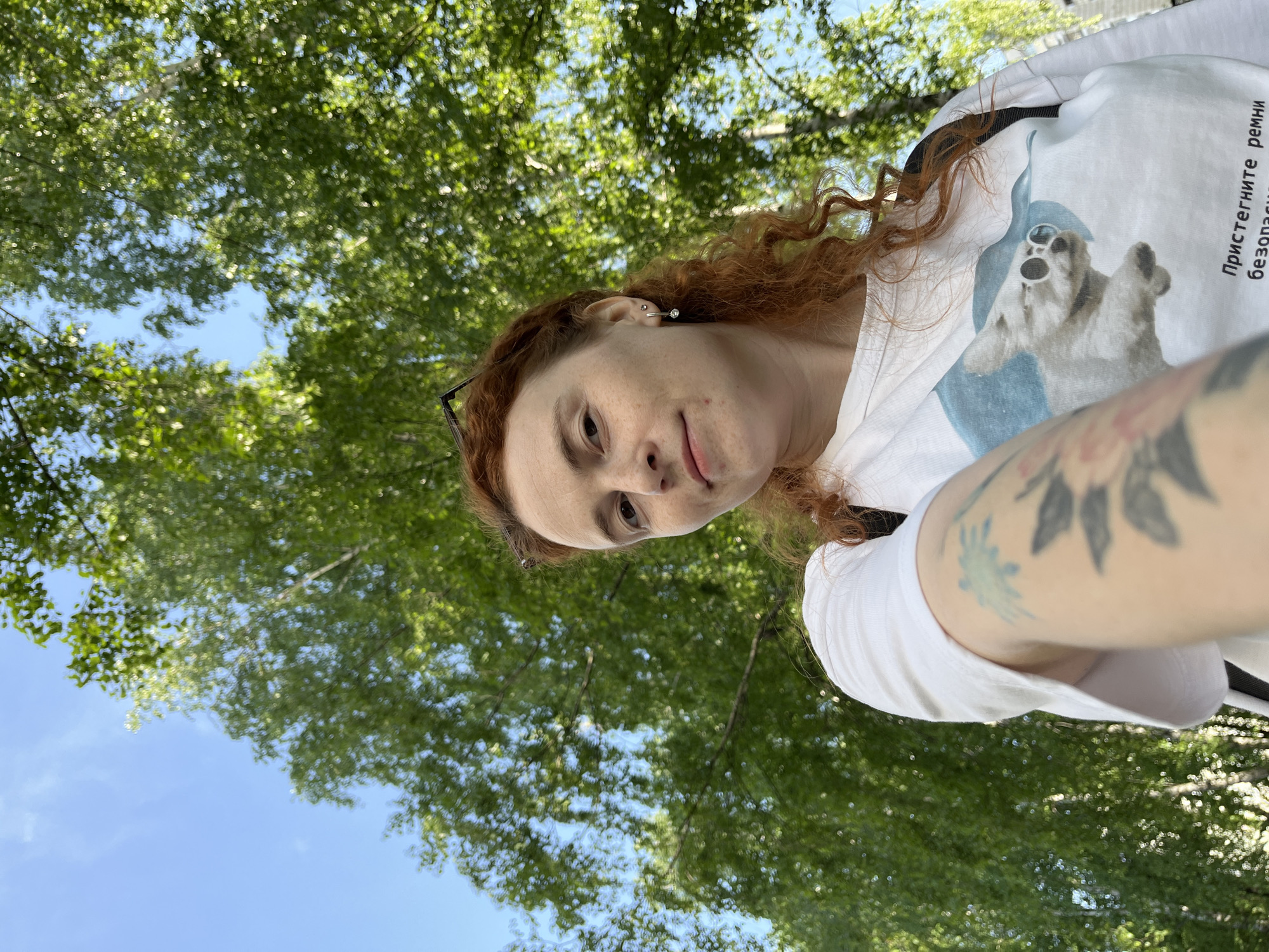 A selfie taken outdoors in what appears to be a park. A woman with red, curly hair is the main subject, partially in focus. She is wearing a white t-shirt with a cartoon bear printed on it. A tattoo is visible on her upper arm. Behind the woman, a canopy of green leaves suggests she is standing under trees. Patches of blue sky can be seen through the leaves.
The location cannot be determined to be the city of Chaikovsky, Russia based on the visual information alone.