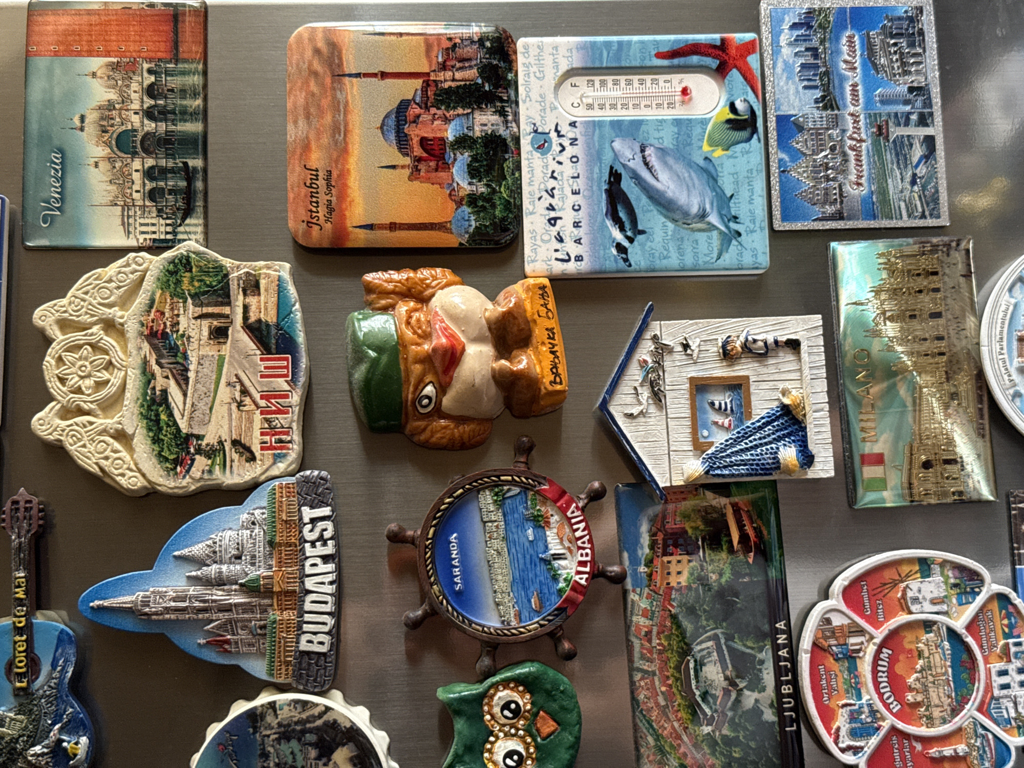 The image shows a collection of refrigerator magnets depicting various European cities and landmarks. The magnets are arranged on a metallic surface, likely a refrigerator door. The cities represented include Budapest, Venezia, Saranda (Albania), Istanbul, Ljubljana, Bodrum (Turkey), Milan (Italy), and Frankfurt am Main (Germany). There are also magnets with designs of a guitar, an owl, a ship's wheel, a cartoonish bird-like character, a beach house, and a thermometer with marine life. The lighting is bright, suggesting an indoor setting. There are no people or animals present in the image. The specific location in the city of Pleven, Bulgaria, is not visible in the image.