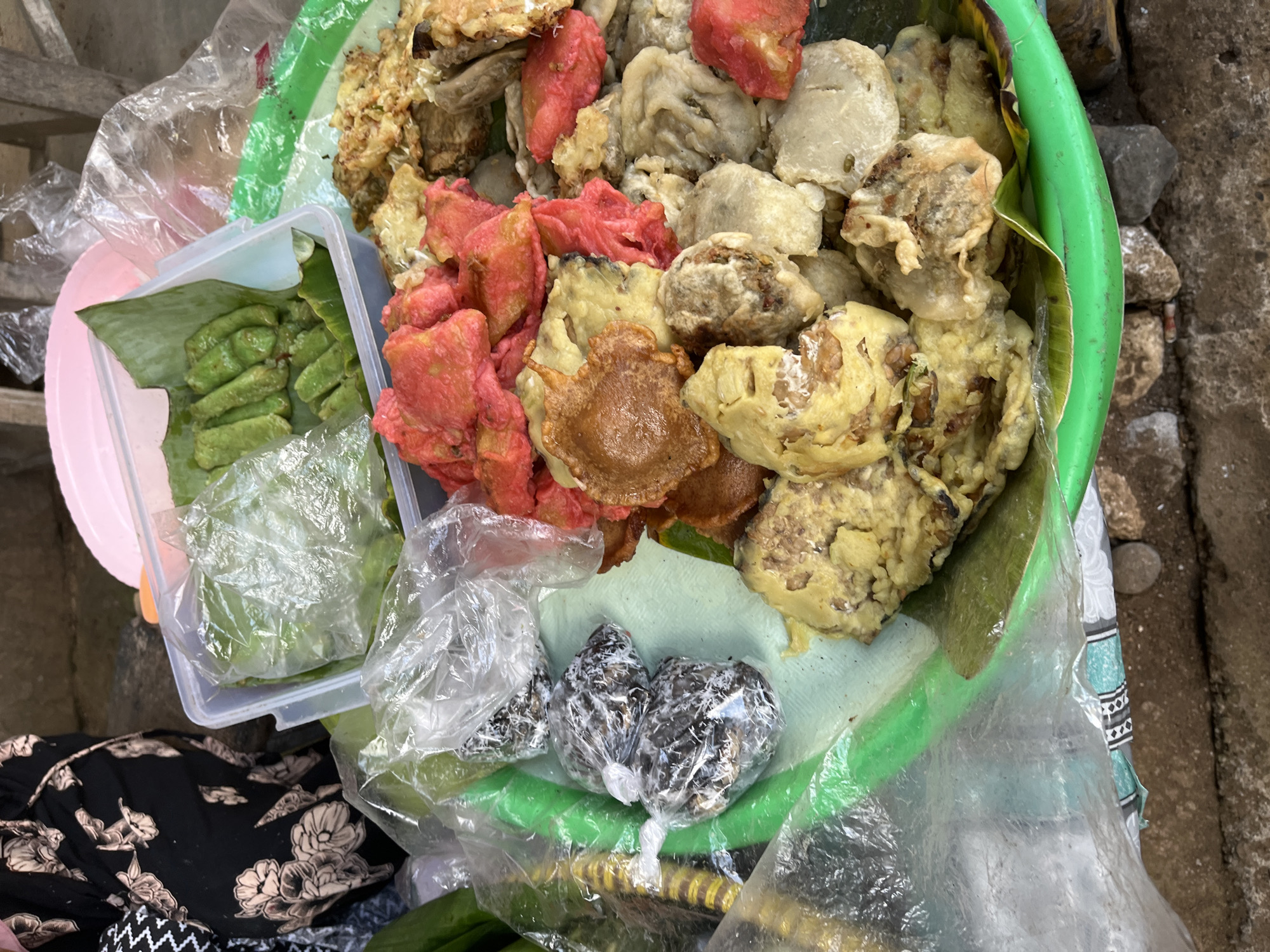 A close-up, overhead shot captures a green plastic tray filled with various Indonesian snacks. The tray, resting on a rough, grey surface, is partially obscured by a clear plastic sheet. To the left of the tray, a person wearing a black dress with a floral print is visible.
The tray's contents include:
* **Green snacks:** A clear plastic container holds several small, green, rectangular snacks, possibly wrapped in banana leaves.
* **Red and golden-brown fried items:** A large portion of the tray is covered with a mix of fried snacks. Some are bright red, while others are golden-brown and appear to be pieces of battered tempeh or other fried items.
* **Wrapped dark items:** To the left of the fried snacks, three small, dark items are individually wrapped in clear plastic, secured with white ties. Their contents are not clearly discernible.
* **Banana leaves:** Some of the snacks are presented on or wrapped in banana leaves.
* **Green cylindrical objects:** Partially visible on the far left are some bright green, cylindrical objects, possibly vegetables or another type of snack.
The overall impression is that of a street food vendor's display in Tambakreja, Indonesia, showcasing a variety of popular local snacks. The lighting suggests an outdoor environment, possibly a market or roadside stall.