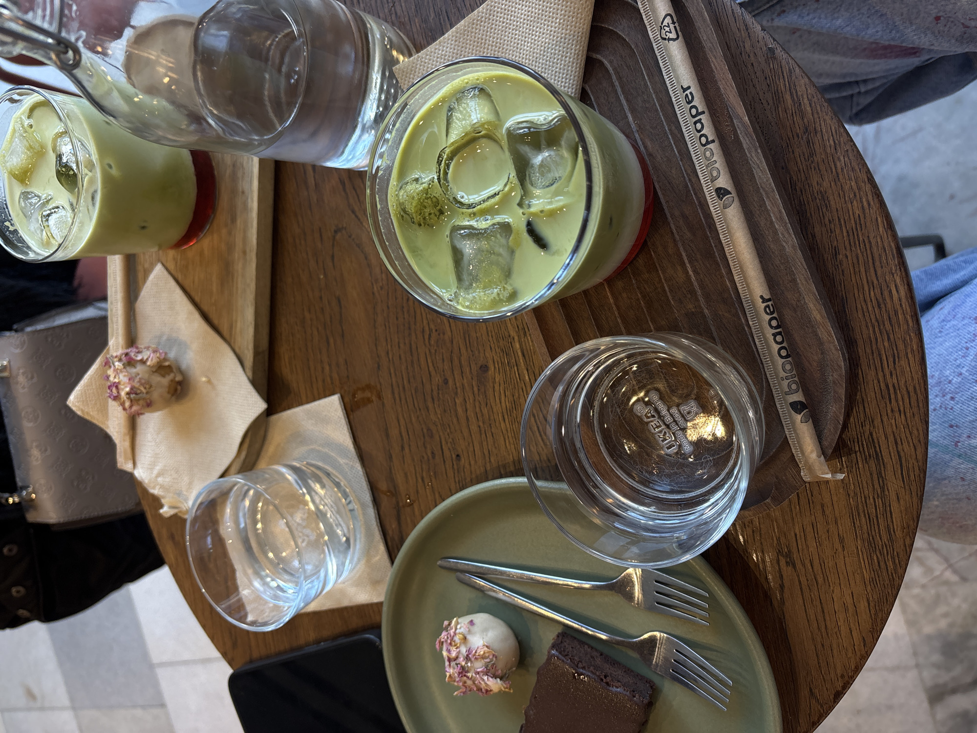 An overhead shot captures a round wooden table at a café, likely in Varna, Bulgaria. On the table is a glass of green-colored matcha latte with ice, a glass of water, and a "biopaper" straw. Another wooden plank holds a matcha drink in a glass and another empty glass. A small plate contains a white treat decorated with rose petals and a piece of chocolate brownie, along with two small forks. A glass of water is placed above this plate. A smartphone is at the edge of the frame. The image also reveals a partial view of the person seated at the table, with blue jeans and a jacket visible. A purse is seen sitting on the floor.