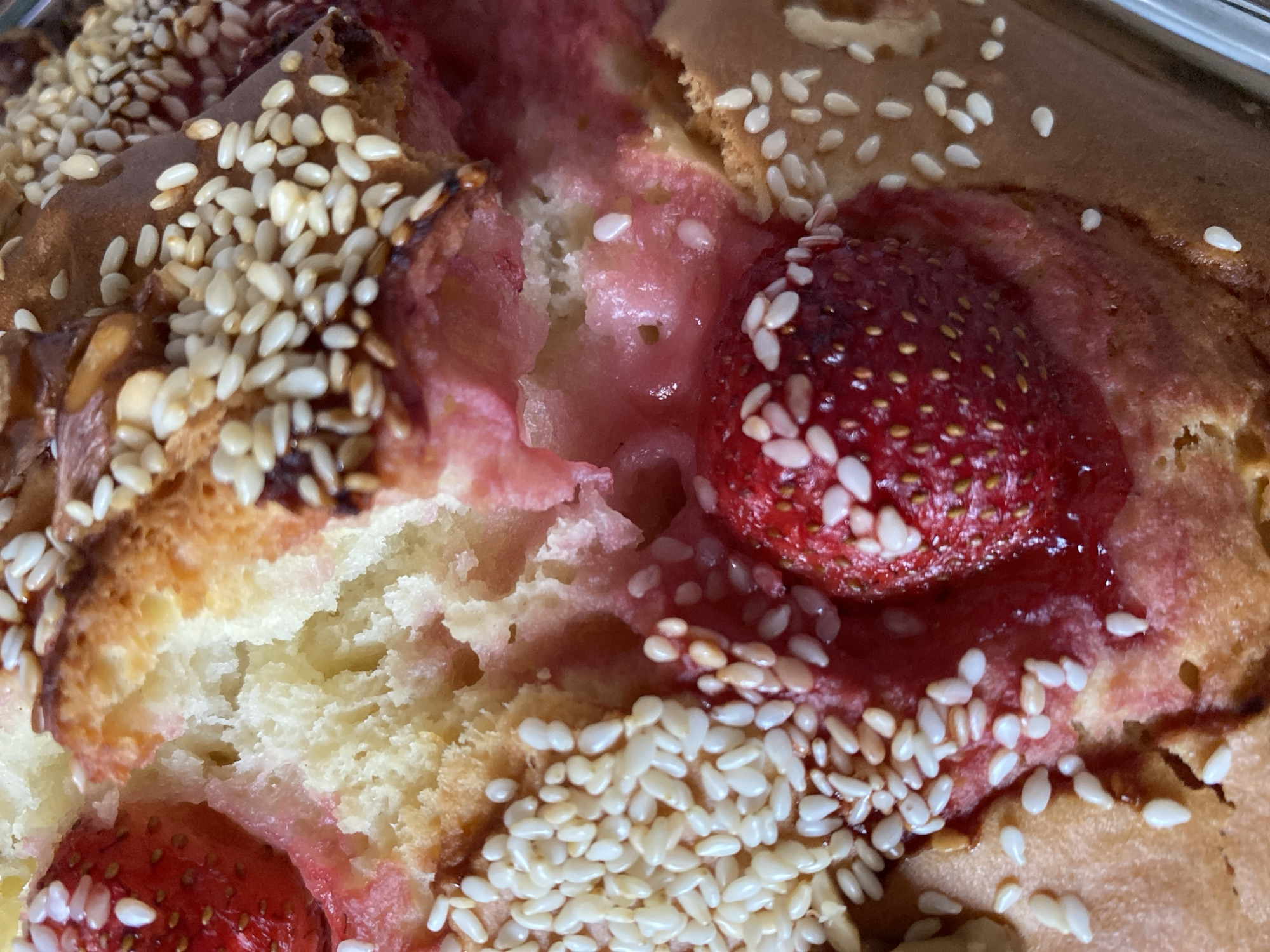 A close-up shot captures a strawberry and sesame seed pastry, revealing its textured crumb and vibrant filling. Whole strawberries are embedded within the golden-brown baked good, their seeds clearly visible. A generous scattering of sesame seeds adorns the crust, some toasted to a darker hue, adding visual interest. The pastry's interior is a soft, pale yellow, interspersed with streaks of pink strawberry filling that oozes from the cracks. The image's sharp focus highlights the intricate details of the pastry's surface, from the individual sesame seeds to the succulent texture of the strawberries. The setting is indoors, with no discernible background elements, suggesting a professional food photography context or a domestic kitchen. No people are present in the frame. The photograph's lighting is bright and even, accentuating the pastry's colors and textures. It is impossible to determine the location of this pastry within the city of Bílina, Czechia, as no contextual information is provided in the image itself.