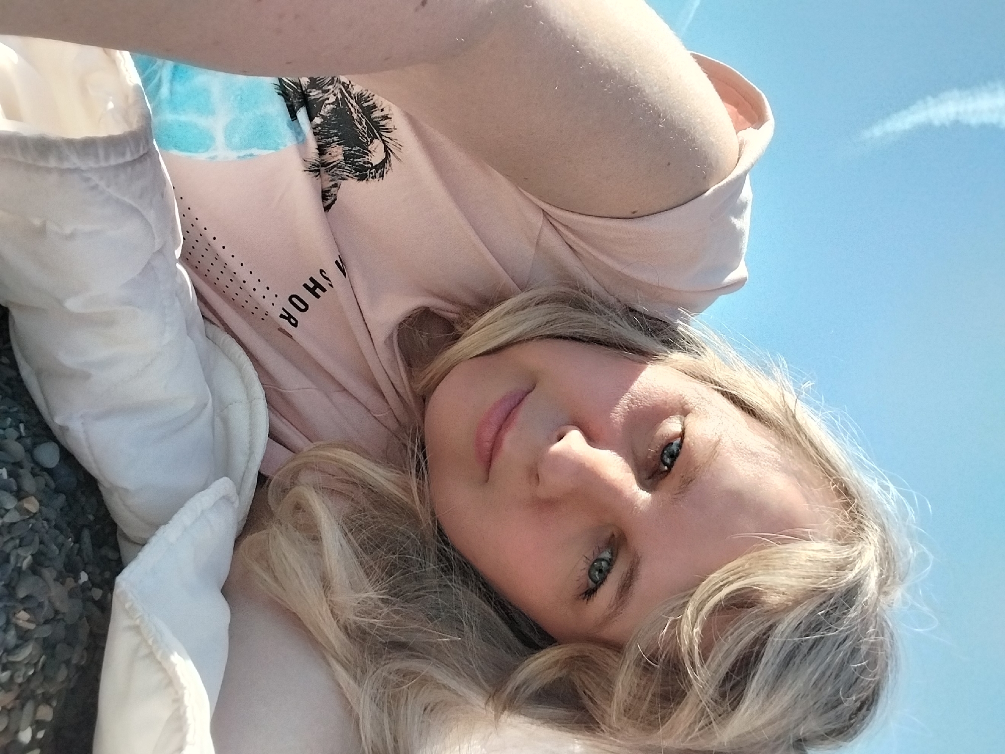 A bright, high-angle portrait captures a young woman with long wavy blonde hair and striking blue-green eyes, looking directly at the camera. She appears to be lying down outdoors, possibly taking a selfie, with a calm and relaxed expression.
She is wearing a light pink t-shirt adorned with a black graphic print of palm trees. Below the graphic, partially visible white text reads "S H O R", suggesting a word like "SHORE" or "SHORT". A light blue and white tie-dye-like pattern is visible near her arm, possibly part of another garment or the t-shirt design. A white, possibly padded, piece of clothing or a blanket is positioned beside her.
The setting appears to be a sunny outdoor location. The background features a clear, bright blue sky with a faint white contrail from an airplane visible in the upper right. In the lower left of the frame, dark grey pebbles or small rocks are visible, indicating a beach or rocky shoreline. The natural lighting suggests it is daytime with clear weather. She has light, natural-looking makeup, enhancing her features.