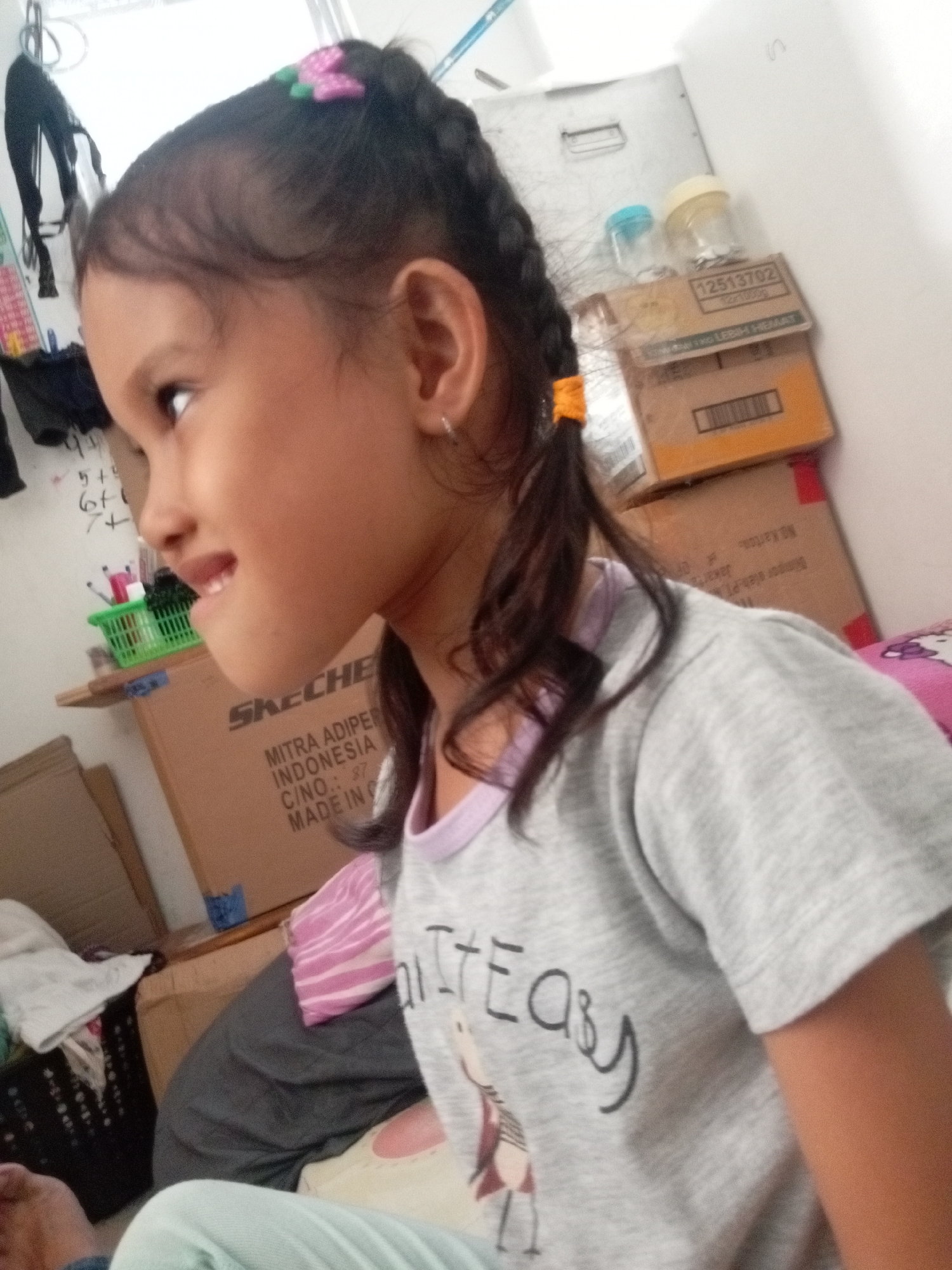 A young girl with braided dark hair, adorned with a pink flower clip, is shown from the chest up, looking to her left. She is wearing a gray t-shirt with a graphic print that reads "Life is Easy". She has a silver earring in her left ear. The background is cluttered with cardboard boxes, a green plastic basket containing pens, and other assorted items. A light-colored wall with some markings is visible behind the boxes. The lighting suggests an indoor setting, possibly a bedroom or storage area. The image appears to be taken in Batam, Indonesia, as indicated by text on one of the cardboard boxes that reads "MITRA ADIPER INDONESIA".