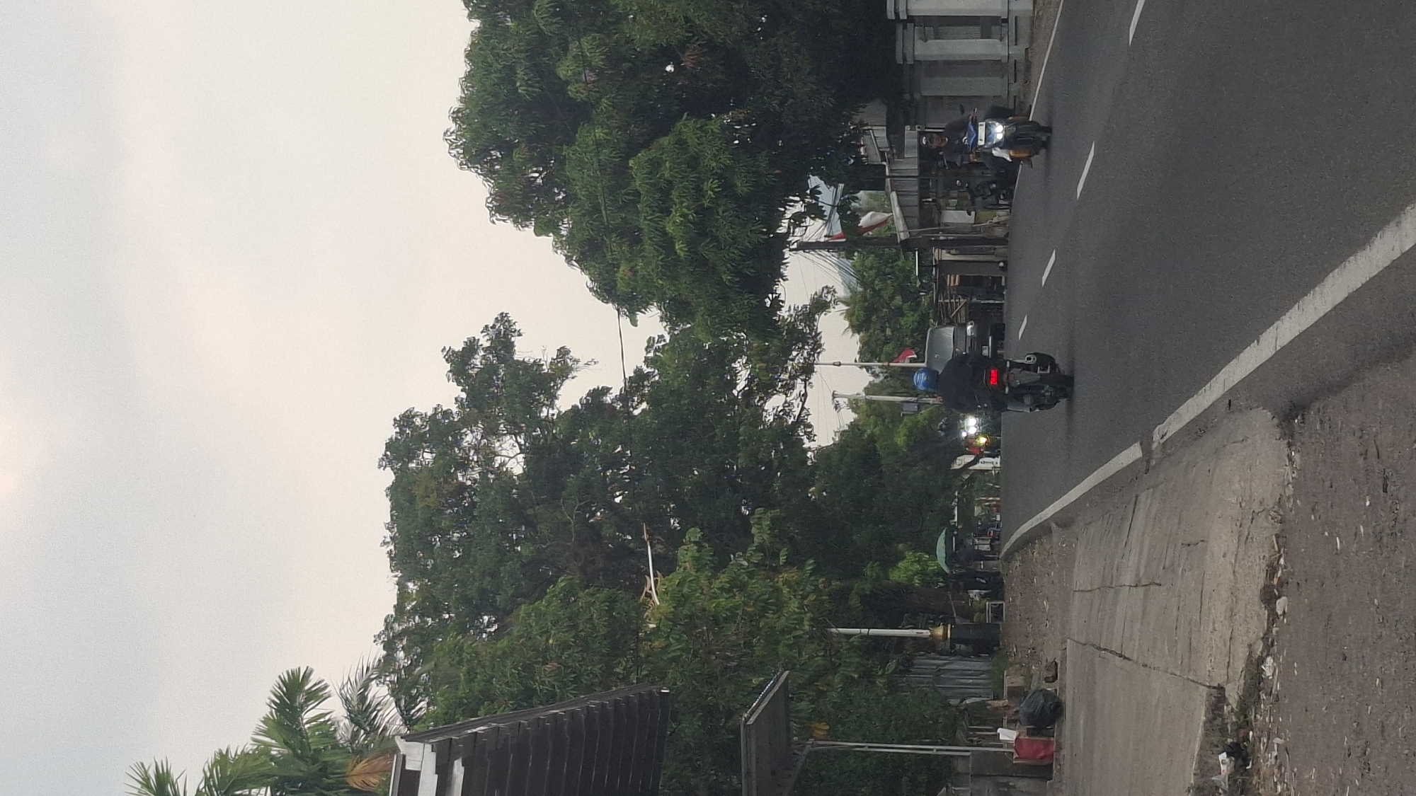 In Sukasari, Indonesia, this image captures a street scene with lush green trees lining a paved road. The sky above is overcast. On the left side of the road, a sidewalk is visible, with some debris on the ground and what appears to be a structure with a sloped roof. Further down the sidewalk, a dark trash bag is placed near the curb.
The road itself is asphalt and has a white line marking the edge. Two motorcycles are traveling on the road. The one in the foreground on the right is ridden by a person wearing a dark jacket and a helmet. The motorcycle further ahead on the left is also being ridden by a person wearing a dark jacket and a blue helmet. A dark-colored van is visible behind this motorcycle. There are also some buildings and structures along the right side of the road, including a white fence. The overall impression is a somewhat overcast day in a semi-urban environment with significant tree cover.
