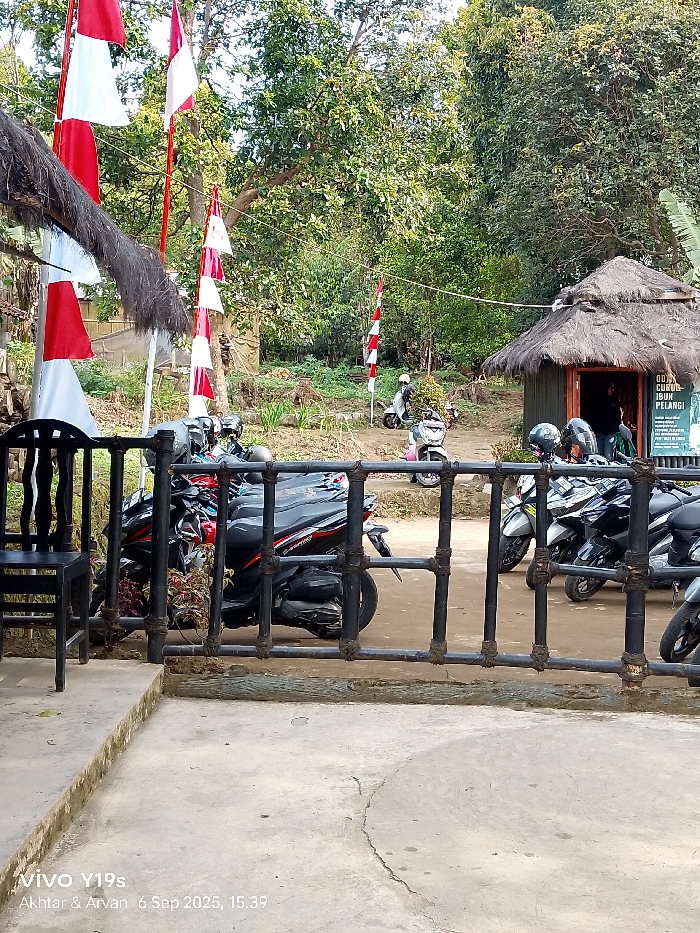 The image displays an outdoor scene in Sukadana, Indonesia, characterized by a parking area and natural foliage. In the foreground, a concrete slab with visible cracks extends from the bottom of the frame, bordering a black metal railing composed of horizontal bars and vertical posts, with the joints wrapped in a dark, fibrous material. A black chair with a slatted back and seat is situated on the concrete to the far left.
Behind the railing, an unpaved dirt path serves as a parking area for at least eight motorbikes, predominantly scooters. A black Honda Vario 160 scooter is parked prominently behind the railing on the left, with additional grey and black scooters to its right, some featuring helmets. Further back on the path, a white scooter and a light pink scooter are visible, each carrying a single individual wearing a helmet; both scooters are oriented towards the right.
To the right midground, a small, dark-colored hut-like building is present, topped with a dense thatched roof made of natural dried plant material. Its walls are constructed from vertical planks, and a dark entrance aperture reveals a dark-clothed figure dimly visible inside. A black sign with white lettering, partially readable as "O OJA CURUG IBUN PELANGI," is affixed to the right side of this structure.
The background is filled with dense green foliage and numerous trees, establishing a natural, wooded environment. Multiple red and white flags, resembling the Indonesian national flag, are displayed vertically on poles. Some flags are positioned on the left near a partial thatched roof structure, and others are distributed further back within the wooded area. The scene is illuminated by natural daylight, consistent with the embedded timestamp of September 6, 2025, at 15:39.