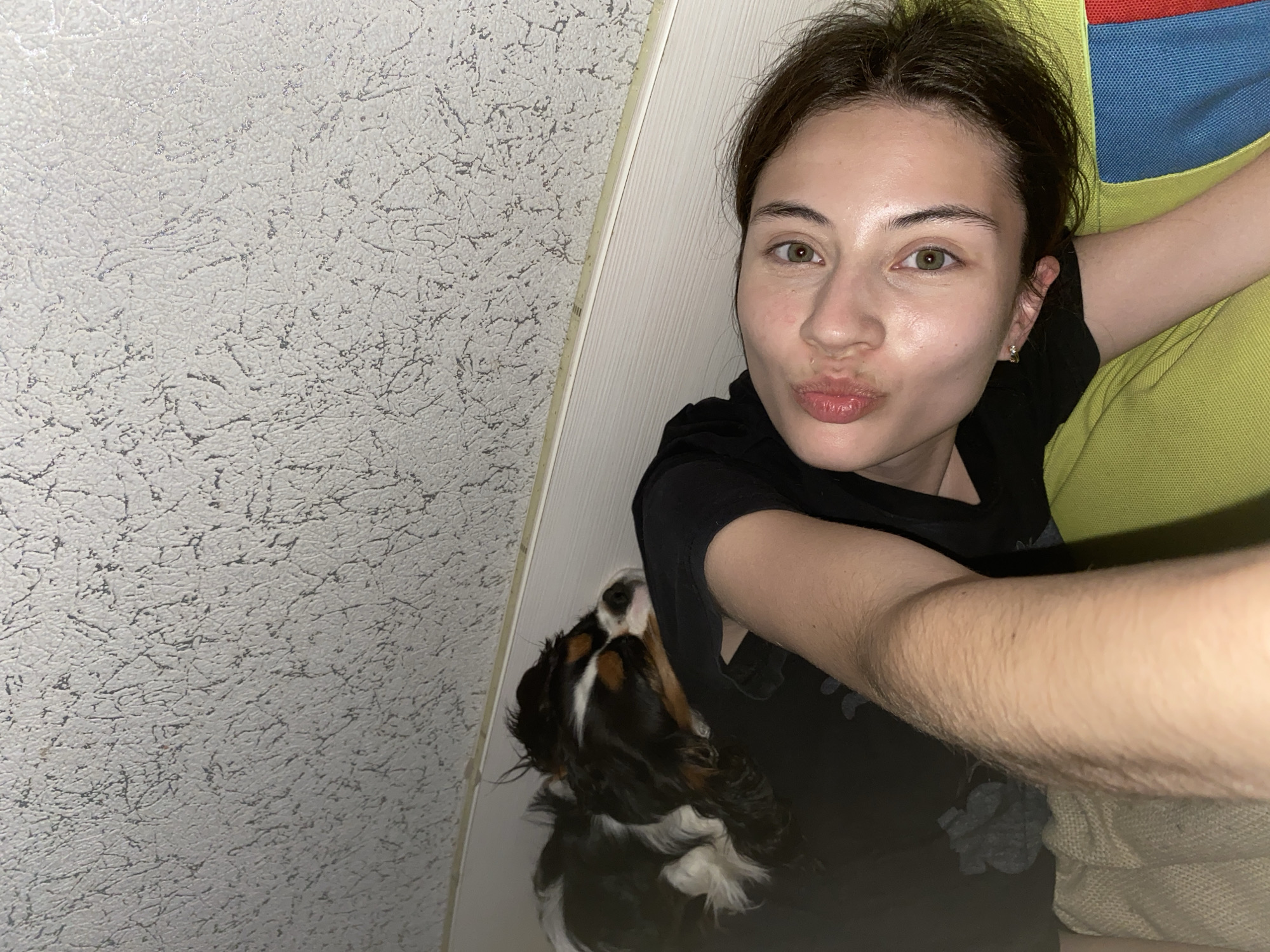 A young woman with green eyes and brown hair is lying down, looking at the camera and puckering her lips. Her arm is extended, as if taking a selfie. To her left, a tri-colored Cavalier King Charles Spaniel is sleeping on her. The background consists of a textured wall with a wooden trim. The scene is indoors, likely a bedroom. There is no indication of the location being in Votkinsk, Russia.