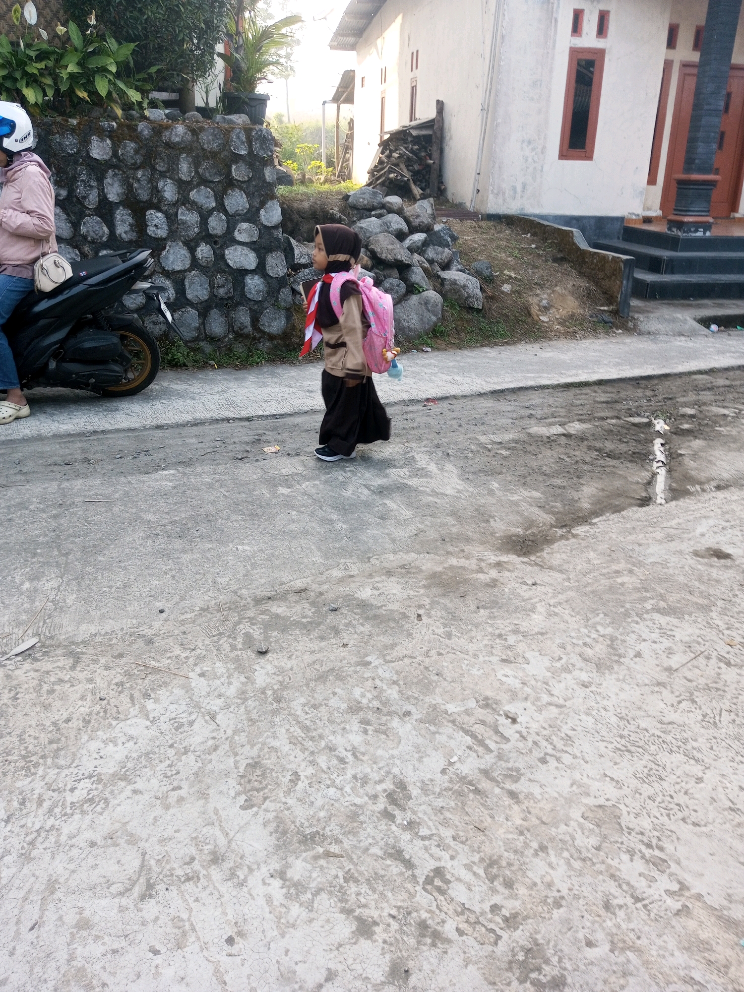 In Krasak, Indonesia, a child wearing a brown hijab and a school uniform walks on a paved street. They are carrying a pink backpack. To the left, a person sits on a motorcycle, wearing a pink jacket and a white helmet. Behind them, a stone wall is visible, bordered by green foliage. A white building with brown accents stands in the background. The ground is a mix of concrete and dirt.