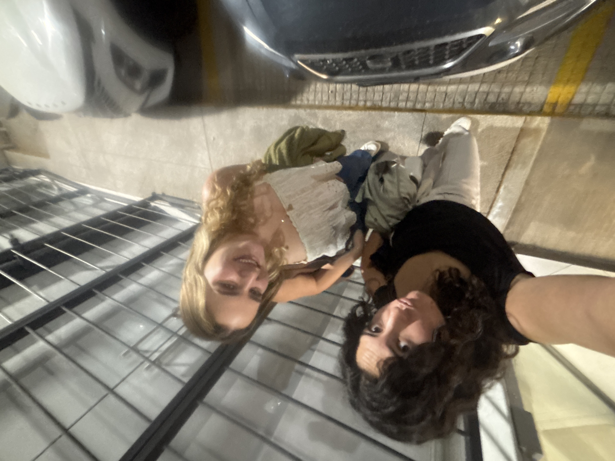 Two young women pose for a selfie taken from a high angle, possibly a rooftop. The woman on the left has long blonde hair and is wearing a white top. The woman on the right has dark curly hair and is wearing a black top. They are standing on or near a metal grid structure, likely part of the building's architecture. Above them, parts of two cars are visible, a white car on the left and a dark car on the right. The ground is paved, and there is a yellow line. There are clothes scattered on the ground. The location is Puebla, Mexico.