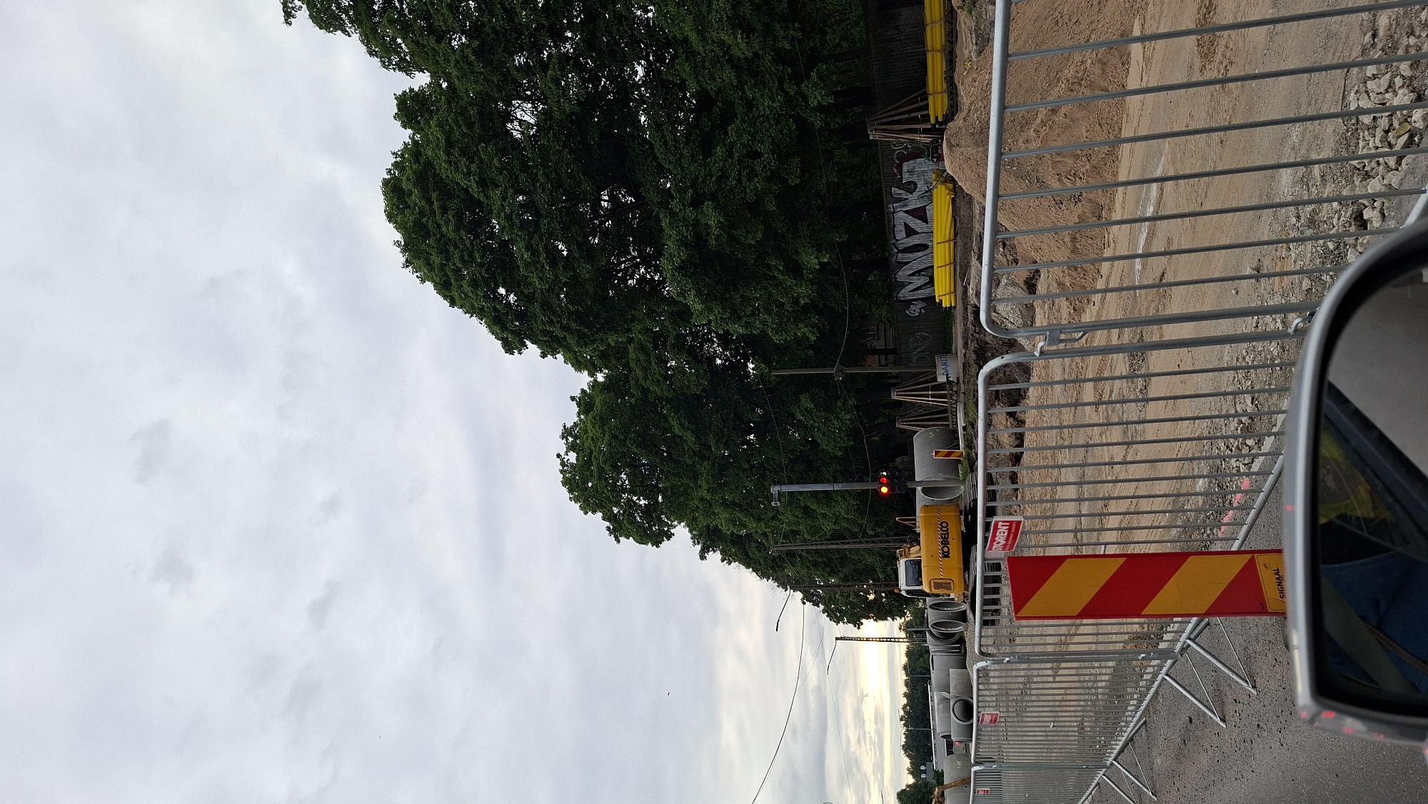 This is a media file taken from a vehicle's perspective, showing a construction site in Tallinn, Estonia. The scene is a road under construction, with temporary metal barriers fencing off the work area. A pile of sand and gravel is visible behind the barriers, with some construction materials and pipes stacked nearby. A yellow Kobelco excavator is parked on the site, and a traffic light displays a red signal. A sign with red and yellow diagonal stripes is placed near the barrier, likely indicating a hazard or a warning. In the background, lush green trees line the street, contrasting with the rough construction environment. The sky is overcast with clouds, suggesting it might be late afternoon or early evening. Some graffiti is visible on a wooden fence behind the trees, adding a touch of urban artistry to the scene. The overall impression is one of ongoing public works, with the elements of nature and urban infrastructure coexisting.