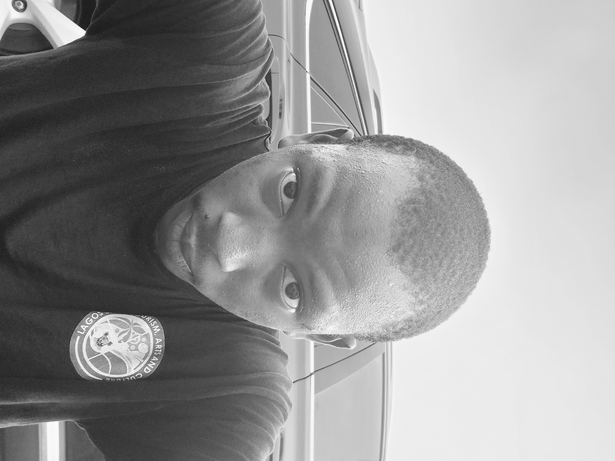 A black and white close-up photo shows a man wearing a t-shirt with the logo "Lagos Tourism, Arts, and Culture". The man appears to be standing near a vehicle. There is no further information regarding a specific location in Lagos, Nigeria.