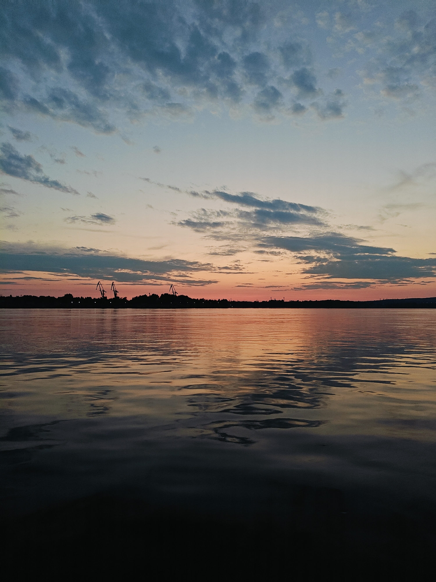 The image depicts a tranquil sunset over the Kama River in Chaikovsky, Russia. The sky transitions from a pale blue at the top to warm hues of orange and pink near the horizon, dotted with scattered clouds. A silhouetted shoreline separates the sky from the water, featuring a line of trees and several industrial cranes. The river's surface reflects the colors of the sky, with gentle ripples creating a dynamic pattern on the water. The lower portion of the image is a dark, almost black expanse, suggesting the approaching night. There are no people or animals visible in the scene.