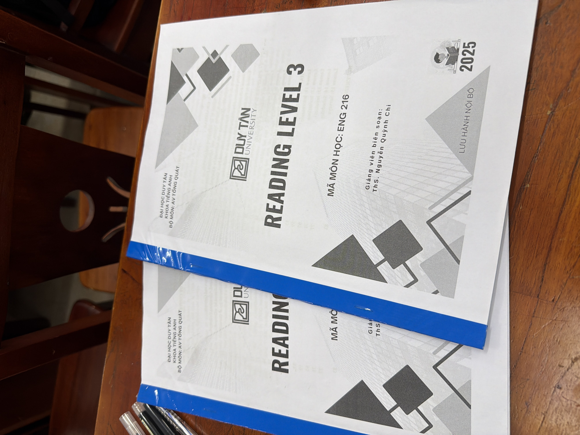 Two blue-bound booklets lie on a wooden table. The top booklet prominently displays "READING LEVEL 3" and the "DUY TAN UNIVERSITY" logo, both in bold print. Beneath, smaller text notes the subject matter (presumably a reading course) and the instructor. The year "2025" is printed in the upper-right corner, along with the words "LƯU HÀNH NỘI BỘ," indicating internal use. The bottom booklet is similarly designed. Both booklets are located at Duy Tan University in Đà Nẵng, Vietnam. There are some pens and pencils on the lower left side. The background has a wooden chair.