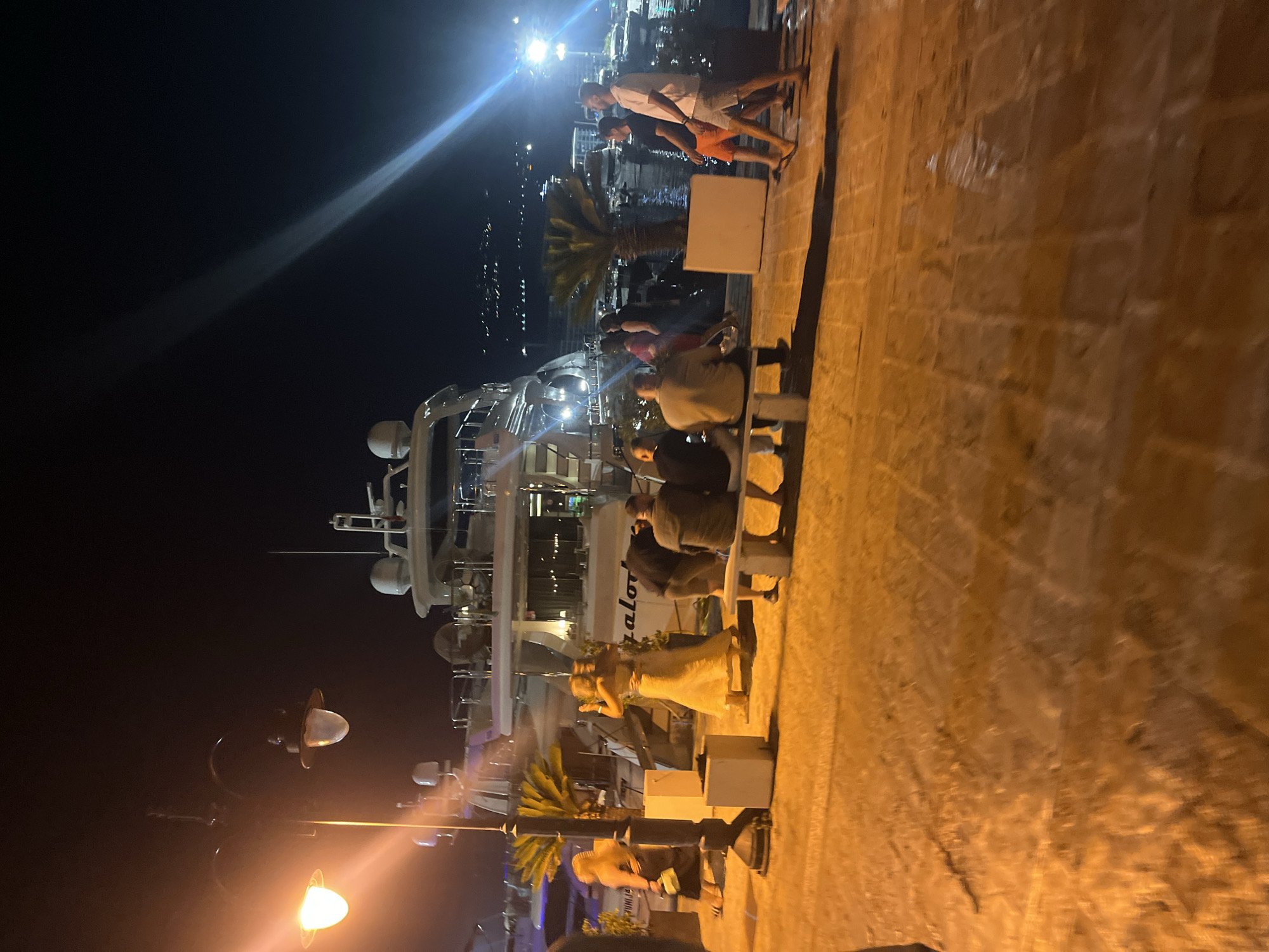 A nighttime scene depicts a waterfront promenade in Budva, Montenegro, with a large yacht docked in the background. Several individuals are present on the promenade. In the foreground, a group of men are seated on a stone bench, facing away from the camera. To the left, two women, one in a long yellow dress and the other in a dark dress, appear to be dancing or posing. Further to the left, another woman in a dark dress walks away from the camera. On the right side of the frame, two men walk towards the right, one wearing orange shorts and a dark t-shirt, the other in a white t-shirt and light-colored shorts. The promenade is paved with cobblestones, illuminated by streetlights that cast long shadows. Palm trees are visible in planters around the yacht. The water of the harbor is dark, reflecting the lights from the shore and boats. The sky is dark, with distant lights visible on a hillside.