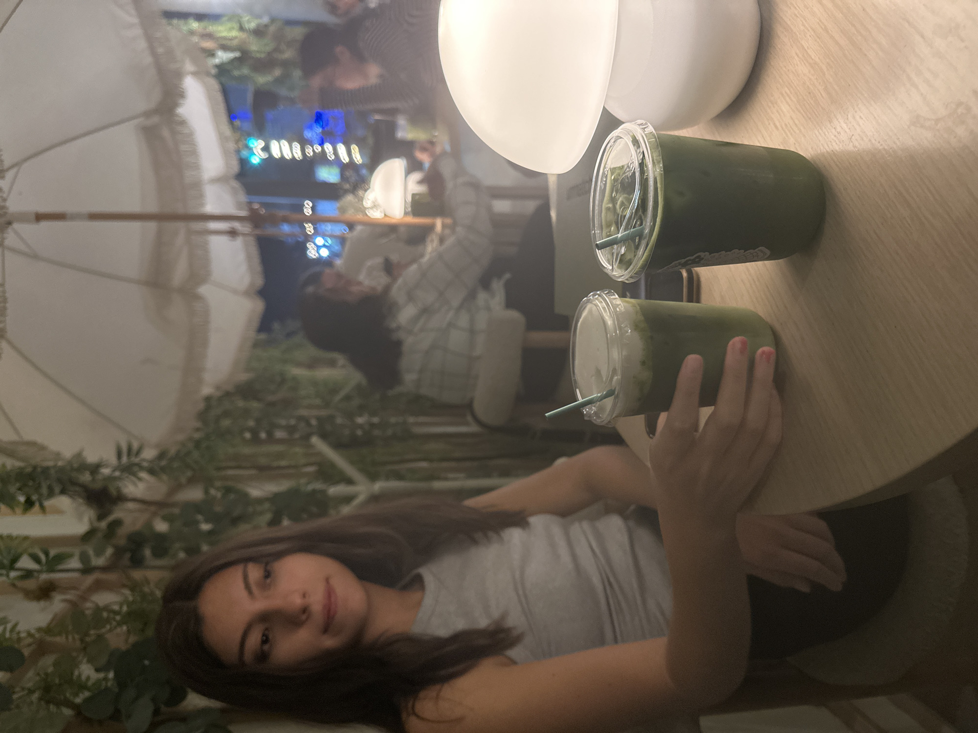 The image shows a person with long brown hair and a gray t-shirt seated at a light wooden table. Two cups of green liquid, likely matcha drinks, are on the table. A hand with red nail polish is near the closer cup. The background includes a white umbrella, greenery, and other people seated. It appears to be an outdoor cafe or seating area. The city of Puebla, Mexico cannot be discerned from the image.