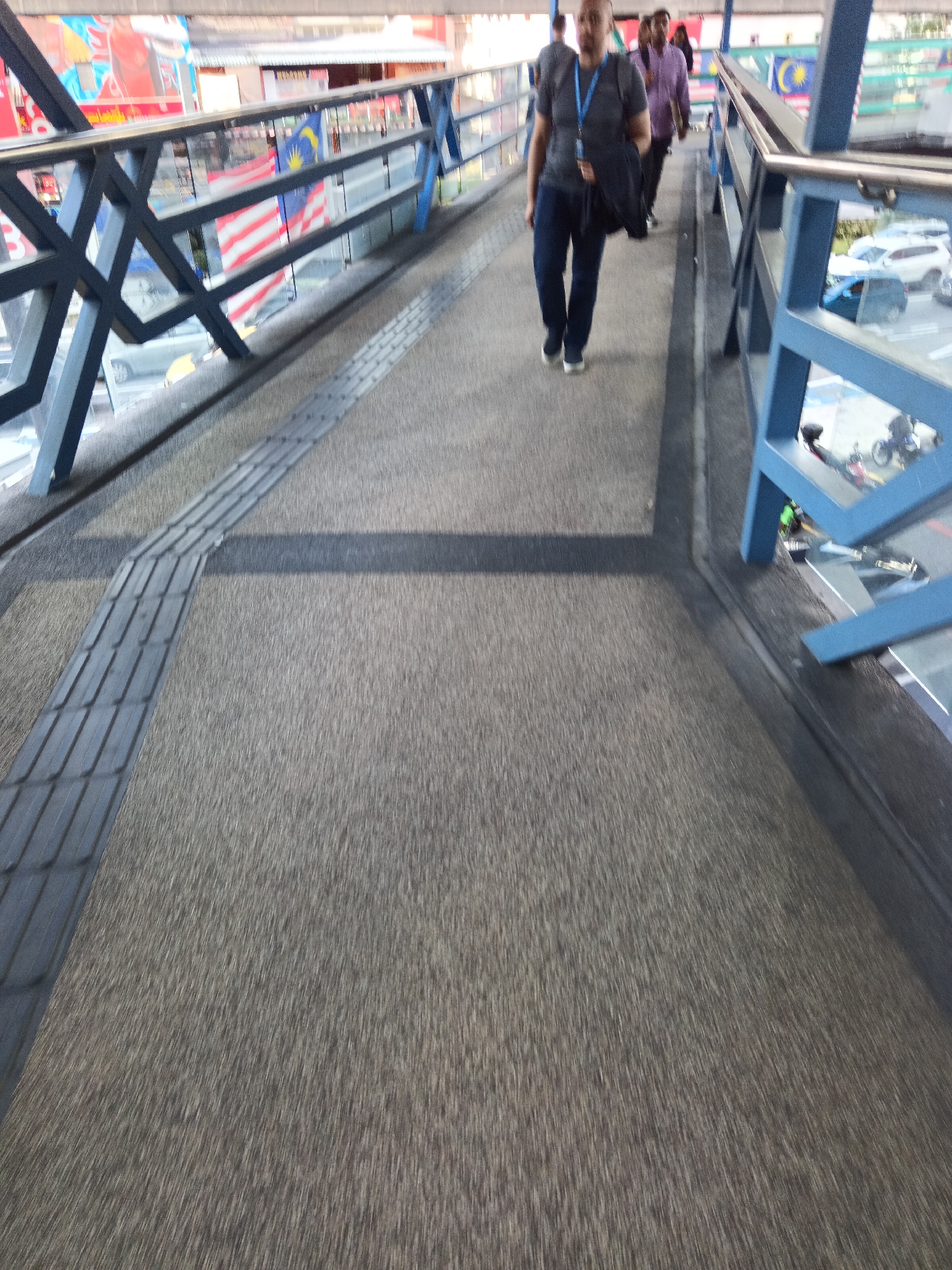 The image depicts a pedestrian walkway, likely an overhead bridge, in Kuala Lumpur, Malaysia. The walkway features a textured grey surface with tactile paving strips for the visually impaired. A black metal railing with blue accents runs along the side, separating the walkway from an area below where vehicles and a motorcycle are visible. Malaysian flags are displayed along the railing, suggesting a public or civic location.
In the foreground and mid-ground, several individuals are walking. The most prominent is a person in a grey t-shirt and dark pants, carrying a black bag over their shoulder. They are walking away from the camera. Behind them, another person in a purple shirt and dark pants is also walking. The background shows a blurred urban environment with buildings and signage, typical of a busy city. The overall impression is a functional pedestrian passage within an urban setting.