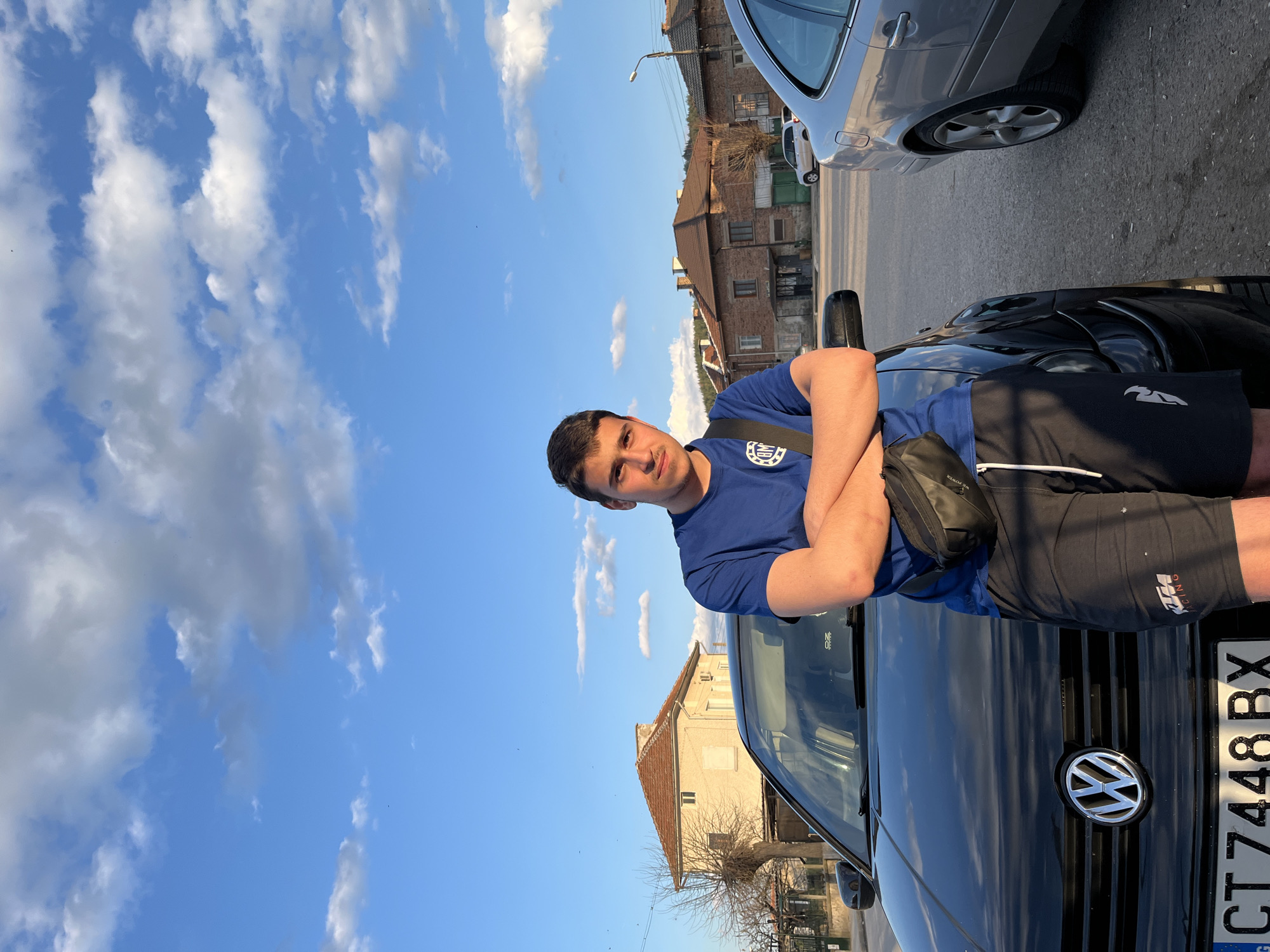 A young man stands next to a black Volkswagen car, parked on a street lined with buildings. He is wearing a blue t-shirt and black shorts and has a black bag across his chest. The car's license plate reads "CT 7448 BX." The background shows houses and a clear blue sky with scattered clouds. The scene is in an urban or residential area, identified as Karavelovo, Bulgaria based on the license plate country code.