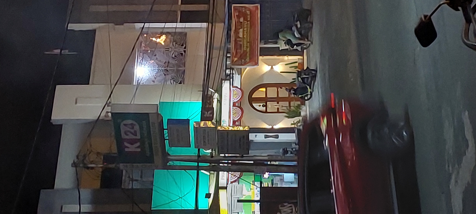 The image depicts a street scene at night in Jayapura, Indonesia. In the foreground, a red car is blurred due to motion, indicating it is moving. Behind the car, there is a sidewalk and a building. The building features a prominent green sign with "K 24" in white letters, suggesting a pharmacy or convenience store, and the text "Apotechir Widana" and "Buka Sampai 24 Jam" below it. To the left of this sign, a large, brightly lit teal-colored panel with a web-like structure is visible.
Further down the street, the building has an arched entrance with wooden doors, illuminated by warm-toned wall lights. Two potted plants and several tall, thin plants are placed on either side of the entrance. Two motorcycles are parked in front of the building. One is a black scooter with a rider wearing a light green outfit seated on it. Another black scooter is parked in front of it. To the right of the arched entrance, a banner with red and white decorative elements is displayed. Above and to the right of the entrance, a window with a decorative frosted glass panel is lit from within, and flames are visible inside, suggesting a fire. Another banner, orange and red with white text that appears to be Indonesian text, is mounted on the building to the far right. Power lines are strung across the upper portion of the image.