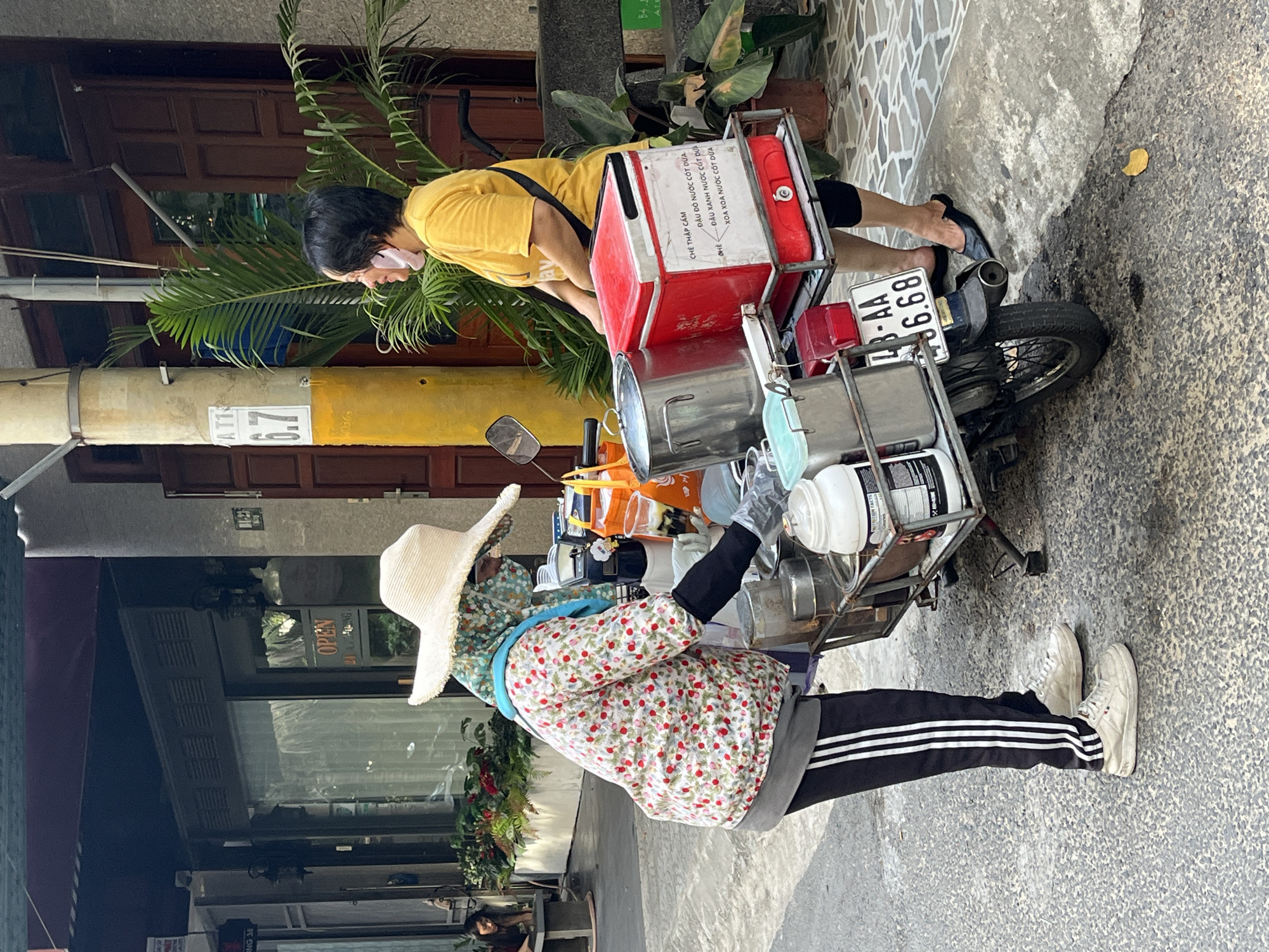 The image captures a vibrant street scene in Đà Nẵng, Vietnam, during what appears to be a bright, sunny day. Two women are interacting around motorbikes, indicating a mobile street food or drink vending operation.
One woman, likely a customer, is dressed in a yellow short-sleeved shirt, black shorts or pants, and flip-flops, wearing a pink face mask. She is leaning over a red insulated container mounted on the back of a black motorbike, possibly making a purchase or collecting an order. The motorbike displays a license plate "43-AA 6.68".
The vendor, positioned on the right, wears a floral long-sleeved shirt for sun protection, black and white striped Adidas-style track pants, white sneakers, a wide-brimmed straw hat, and gloves. She is standing next to a second motorbike, which is elaborately outfitted with a sidecar-like rack holding multiple metal pots, plastic containers, and other vending equipment.
The red insulated container on the customer's side of the bike features a handwritten menu in Vietnamese. Visible items include "CHÈ THẬP CẨM" (Mixed Sweet Soup/Dessert), "CHÈ" (Sweet Soup/Dessert), "ĐẬU ĐỎ NƯỚC CỐT DỪA" (Red Bean with Coconut Milk), "ĐẬU XANH NƯỚC CỐT DỪA" (Mung Bean with Coconut Milk), and "XOA XOA NƯỚC CỐT DỪA" (Grass Jelly with Coconut Milk), suggesting the vendor specializes in traditional Vietnamese sweet soups.
In the background, there are building facades, some with dark wooden elements, and lush green plants. A utility pole is visible, bearing numbers "AT1", "6.7", and "0365". A storefront window shows a partially visible "OPEN" sign. The scene provides a glimpse into the daily life and street food culture characteristic of Vietnam.