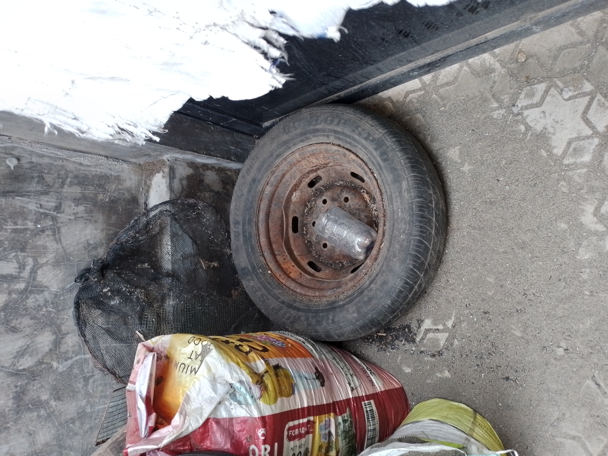 A dirty, rusty tire with a plastic bottle inside lies on a concrete surface in front of a wall in Ngancar, Indonesia. To the left of the tire is a large bag, partially filled with yellow material, with red and white markings. A black mesh bag is behind the tire and the large bag. To the right of the tire, a white, tattered fabric hangs down. The wall behind the objects is rough and grey. The ground is paved with interlocking hexagonal and diamond-shaped concrete blocks, partially covered by dirt and debris.
