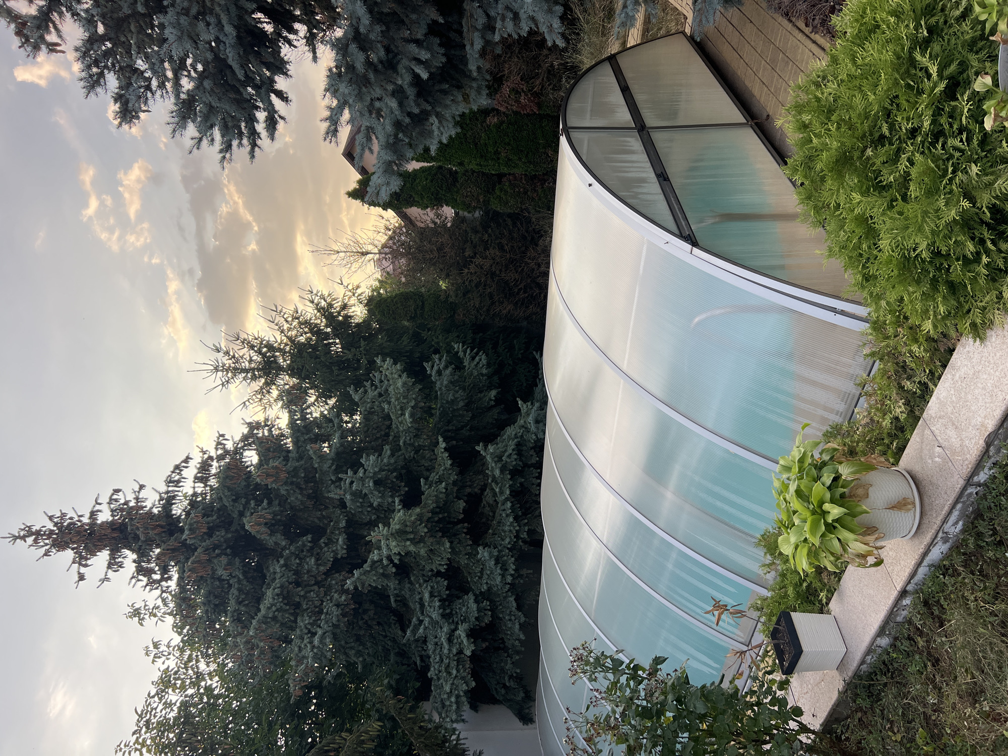 The image shows a backyard with a covered swimming pool. The pool enclosure is made of translucent panels and metal framing. Surrounding the pool are various trees, including evergreen conifers, and lush green bushes. There are also two potted plants on a stone ledge in the foreground. The sky is overcast with some light peeking through, suggesting it might be dusk or dawn. The scene appears to be a residential property in Fontanka, Ukraine, given the context provided. There are no people or animals visible in the image.