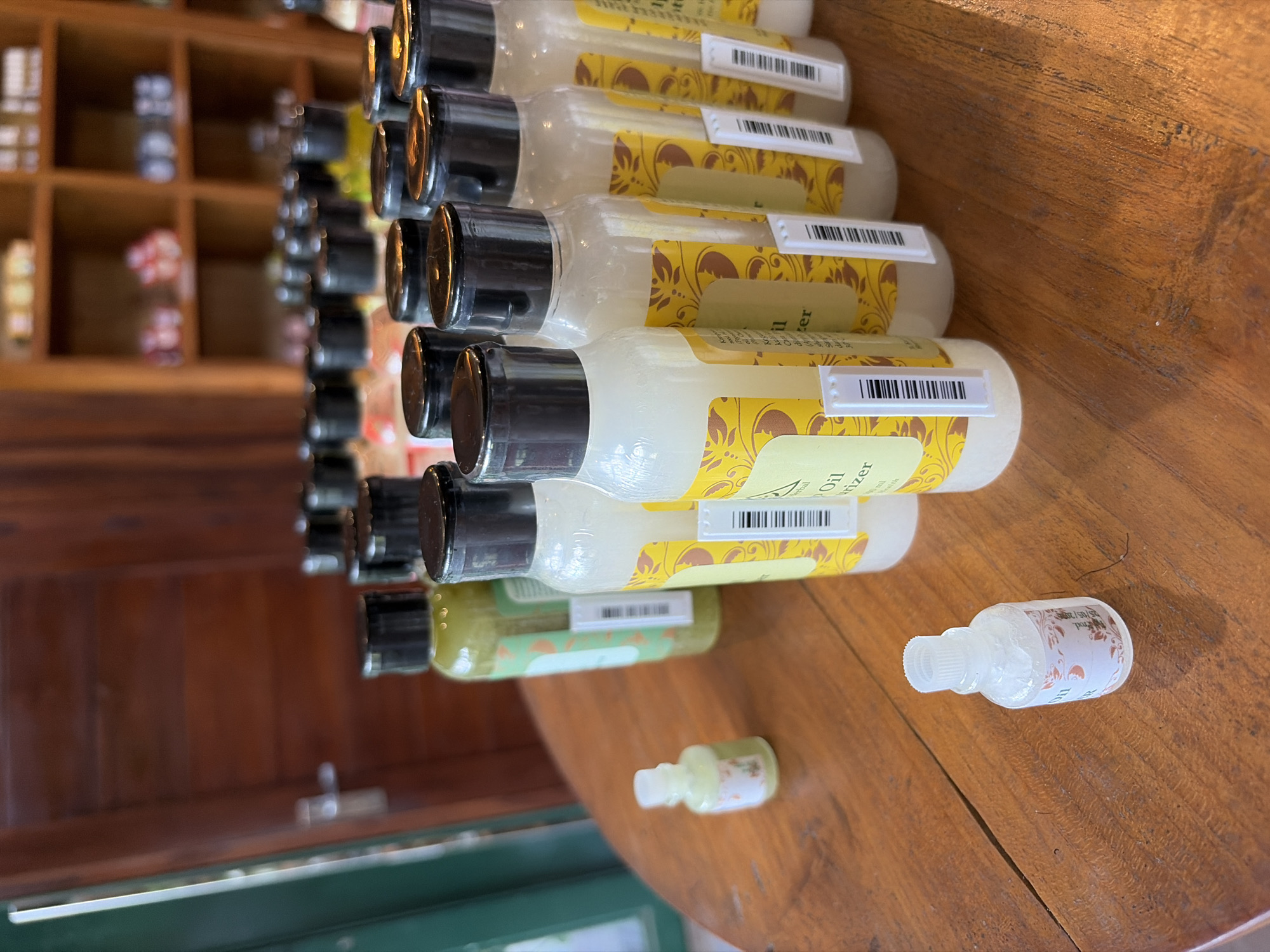 The image shows a wooden table with several bottles of toiletries. In the foreground, a small, clear bottle with a white cap contains a light-colored liquid and a label with floral designs. To the left, a green bottle with a black cap is partially visible. Behind these, a row of clear bottles with yellow labels and black caps are lined up. The labels appear to be in a foreign language. In the background, a wooden shelf unit filled with more bottles and jars is visible, along with a wooden door on the left side of the frame. The overall setting suggests a shop or display area, possibly in Tegallalang, Indonesia, given the wooden furniture and tropical plant motifs on some labels.