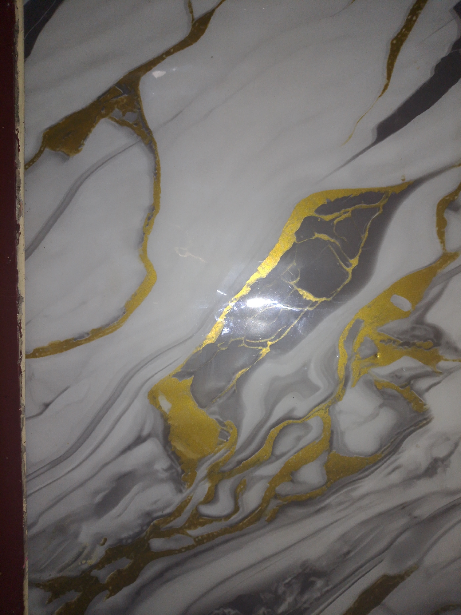 The image shows a close-up of a marbled surface, likely a wall or countertop, with a white base color and veins of gray and gold. The gold veins are irregular and have a metallic sheen, suggesting they are either painted on or made of a metallic material. The gray veins are translucent and blend into the white base. The overall effect is luxurious and modern. The image appears to be taken in a residential or commercial setting within Mukapayung, Indonesia, though no specific landmarks or environmental details are visible to confirm this location. The lighting suggests an indoor environment, possibly with artificial light creating reflections on the glossy surface. There are no people or objects present in the frame besides the marbled material itself.