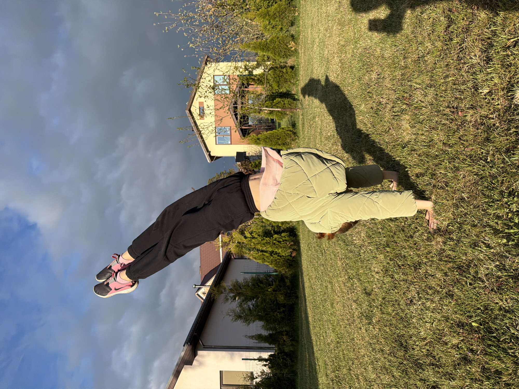 A person in black pants, pink shoes, and a light green quilted jacket is performing a handstand on a grassy lawn. A shadow is cast on the grass behind them. In the background are two houses and a partly cloudy sky. The specific location in Dreiliņi, Latvia is not determinable from the image.