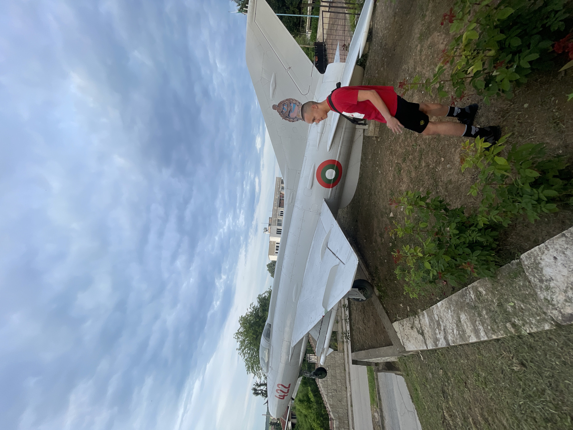 A waist-up shot shows a person in a red shirt and black shorts, who is looking at an airplane, which is tilted to the right, behind them. The plane is white with a red and green target symbol and the number "422" on it. The sky above the plane is cloudy. The environment surrounding the plane includes green vegetation, a gray stone wall, and a street, suggesting the plane is displayed in an outdoor urban area in the city of Unknown, Unknown.