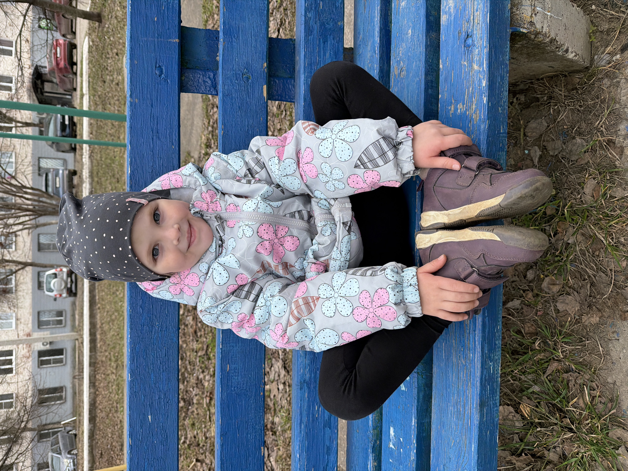 A young child sits cross-legged on a bright blue wooden bench. The child is wearing a gray and black polka-dot hat, a gray jacket with a pattern of blue and pink flowers, and black leggings. The child's feet are wearing brown shoes. The child is holding their shoes. The background is a mix of grass, a building, and trees. The image was likely taken in an urban area, as evidenced by the building in the background. The location is identified as Votkinsk, Russia.