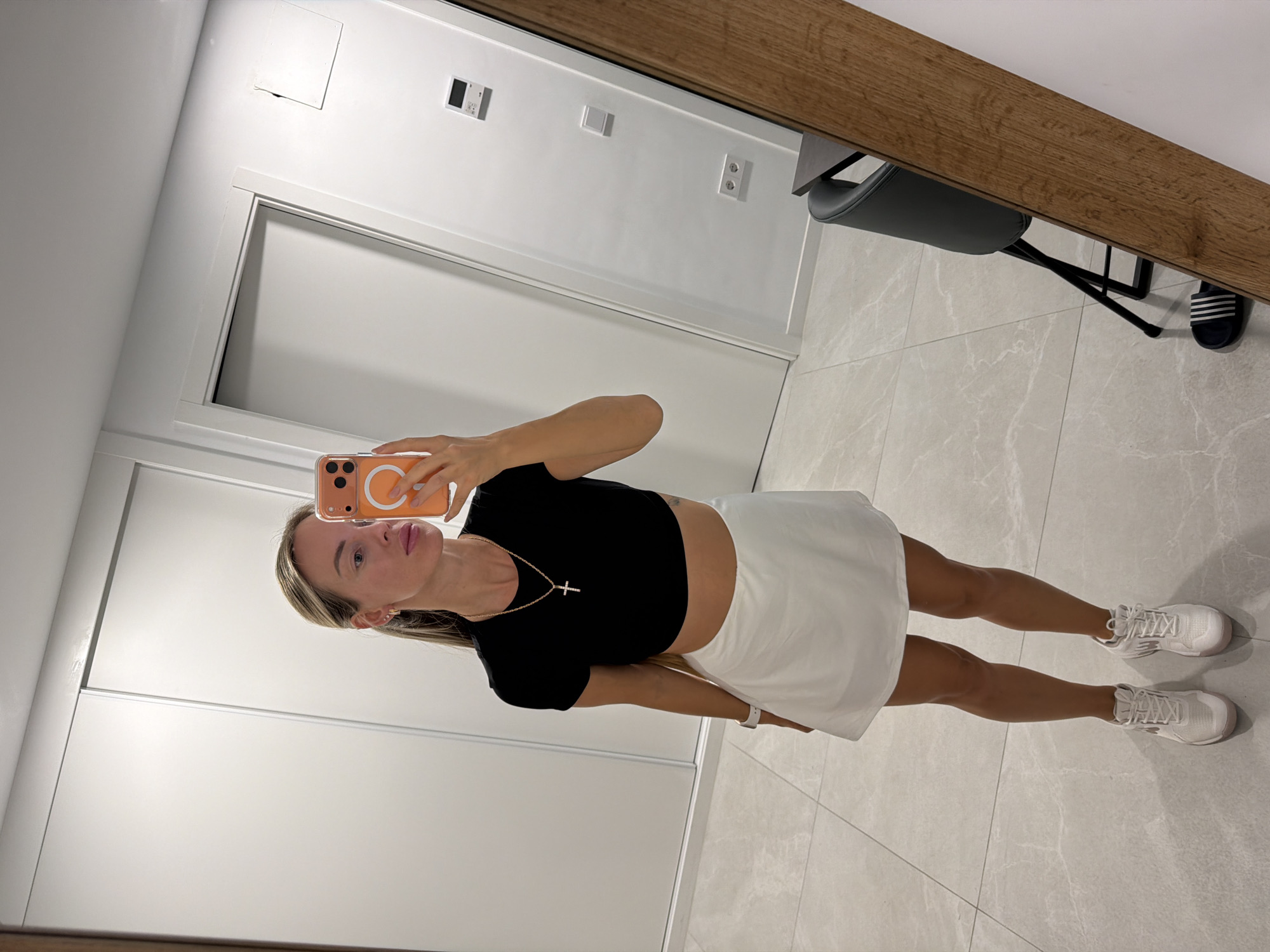 A full-body, mirror selfie of a Caucasian woman in her late 20s or early 30s, standing in a modern, minimalist interior. She has blonde hair pulled back into a ponytail, is wearing a black crop top, a white mini-skirt, and white athletic shoes. She is holding an orange-cased smartphone up to take the photo. Behind her, a large, white sliding closet door is visible, along with a white-paneled door and a wall with light switches and electrical outlets. The floor is tiled with large, light gray marble-patterned tiles. To the right, a portion of a wooden-framed mirror and a dark gray stool with black legs are visible. This is likely an apartment or hotel room in Finestrat, Spain.
