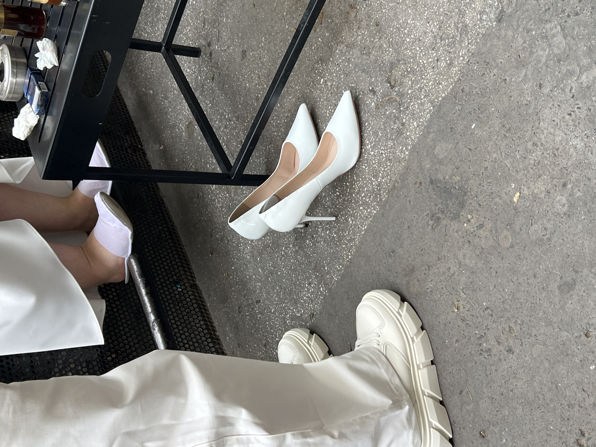 The scene is an outdoor setting in Belgrade, Serbia. The ground is a concrete surface, showing wear and tear. A person wearing white wide-leg trousers and white platform sneakers stands in the foreground. Next to the person, a pair of white high-heeled pumps lie on the ground. Nearby, there is a black metal table with a tray on top, containing a mix of items, including a metal container and bottles. A person's leg wearing a short white dress and white socks is visible near the table. The overall impression is that of someone transitioning from heels to more comfortable shoes in an urban environment.