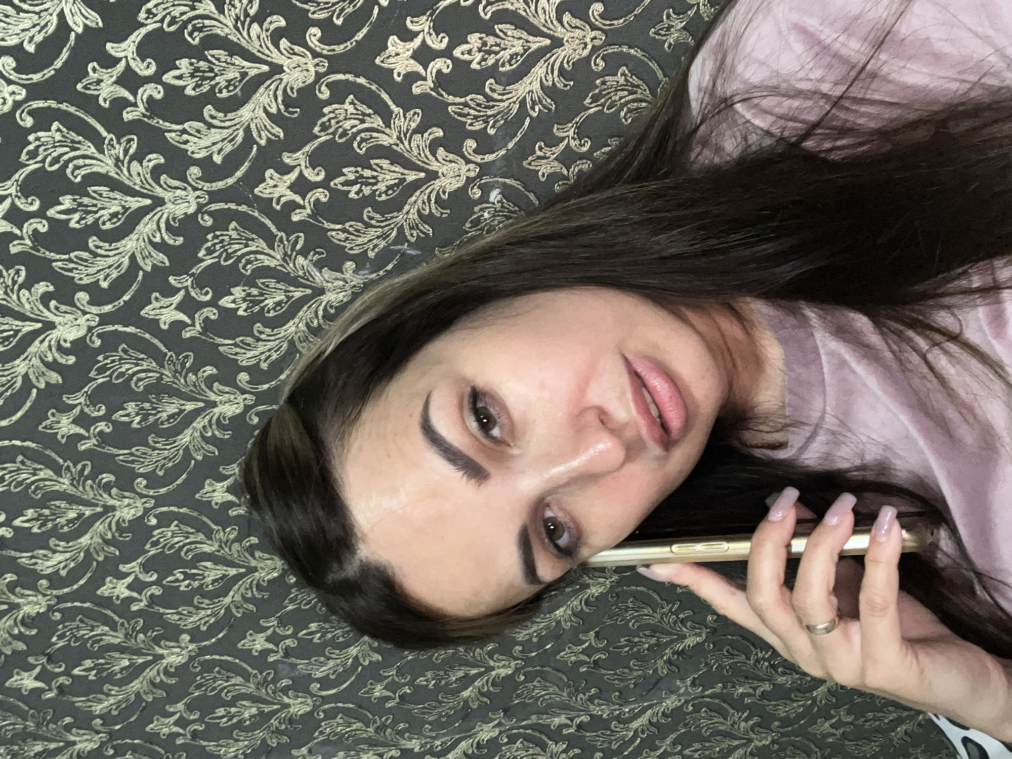 A close-up, low-angle selfie shows a woman holding a gold smartphone to her left ear, in what appears to be a room with ornate wallpaper. The woman has long, dark brown hair, prominent eyebrows, and light brown eyes. She is wearing a pale pink t-shirt. Her left hand, visible in the foreground, has long, light purple fingernails and a thin silver band on her ring finger. The wallpaper behind her is a dark grey with a repeating damask-like pattern in a muted gold color. The lighting suggests it is indoors, with soft light illuminating her face.