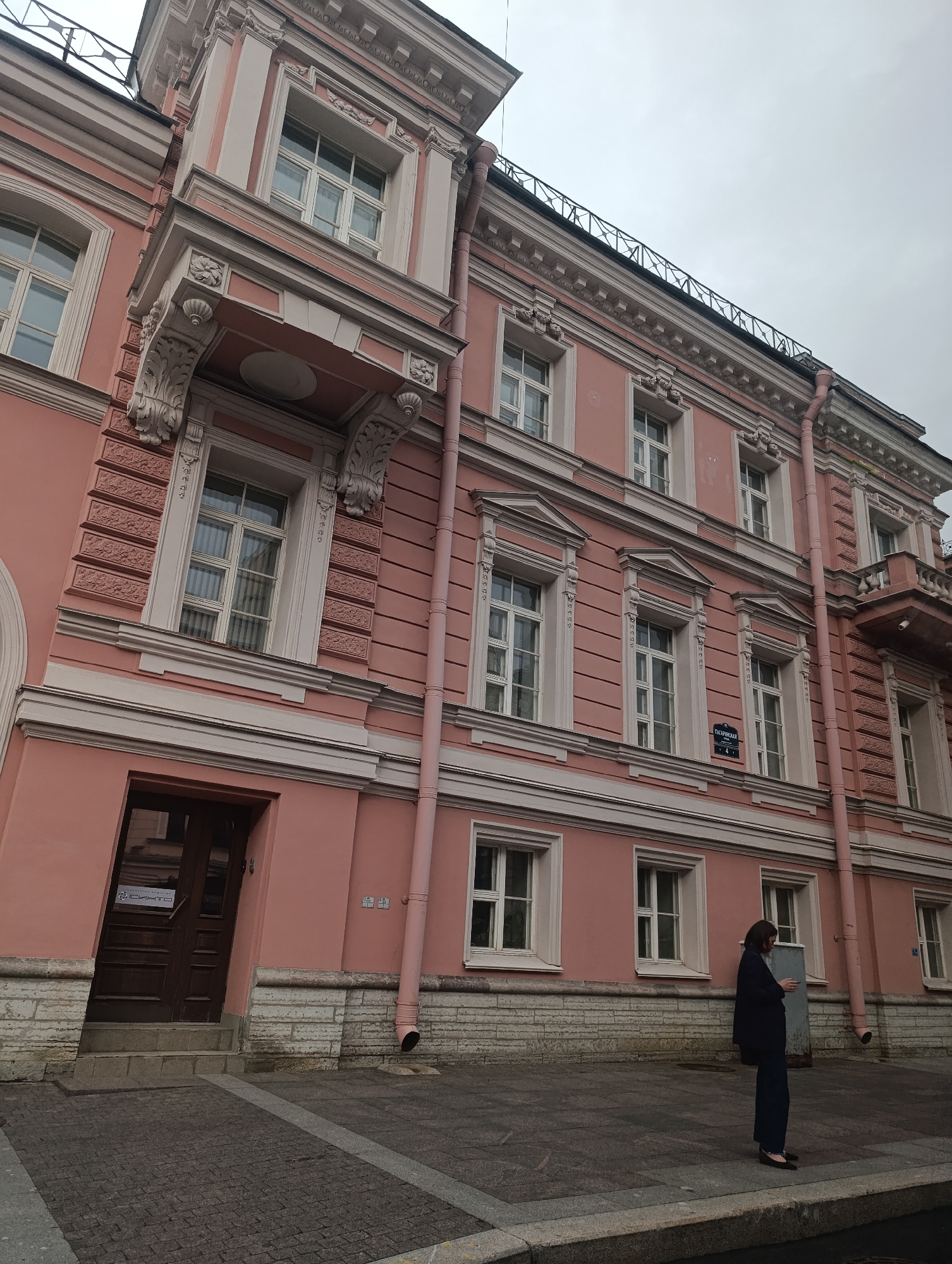 The image shows a woman standing on a sidewalk in front of a large, pink, multi-story building. The building has a classical architectural style with ornate decorations around the windows and a detailed cornice. There are multiple windows on each floor, some with intricate white frames. A dark brown wooden door is visible on the ground floor to the left, with a ramp leading up to it and steps on the side. The sidewalk is paved with cobblestones and there are some granite elements. A pink downspout runs vertically along the side of the building. In the background, the sky is overcast, suggesting a cloudy day. The woman is wearing a dark blue jacket and dark pants, and appears to be holding something in her hands. The overall atmosphere is calm and somewhat subdued.
Visible text includes street signs on the building, one of which reads "ТАВРИЧЕСКАЯ" (Tavricheskaya). There is also text on a sign on the door, but it is not clearly legible. The scene is characteristic of Saint Petersburg, Russia, with its grand architecture and often overcast skies.