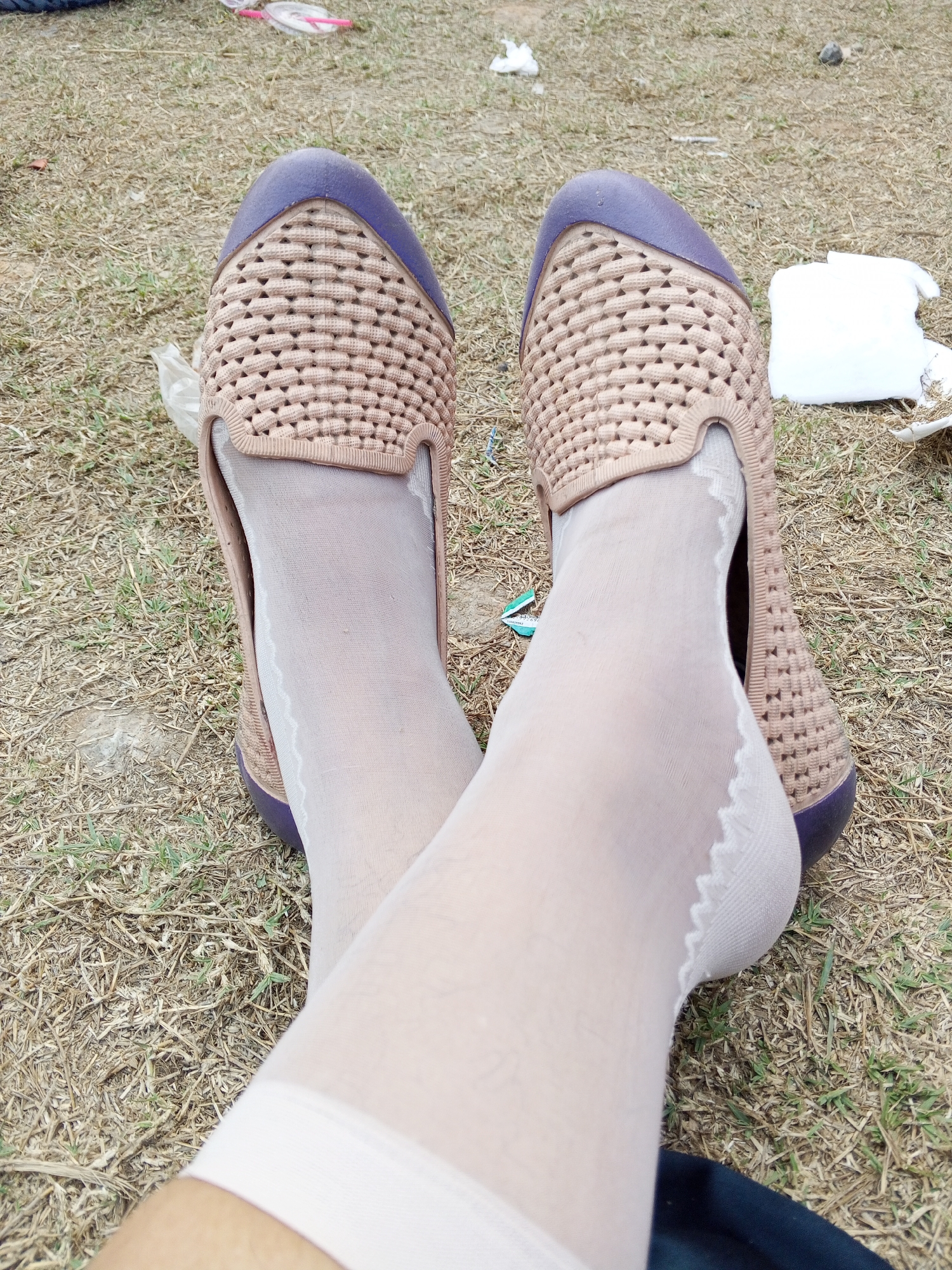 A close-up, eye-level, overhead shot focuses on a person's legs and feet, which are crossed. They are wearing light beige, woven slip-on shoes with purple toe caps and white sheer stockings that have a scalloped trim at the top. The legs and feet are resting on dry grass and soil, with some scattered debris such as plastic wrappers and a white cloth in the background. The scene appears to be outdoors, possibly in a park or open field. No specific location in Ranjeng, Indonesia is identifiable from the image.