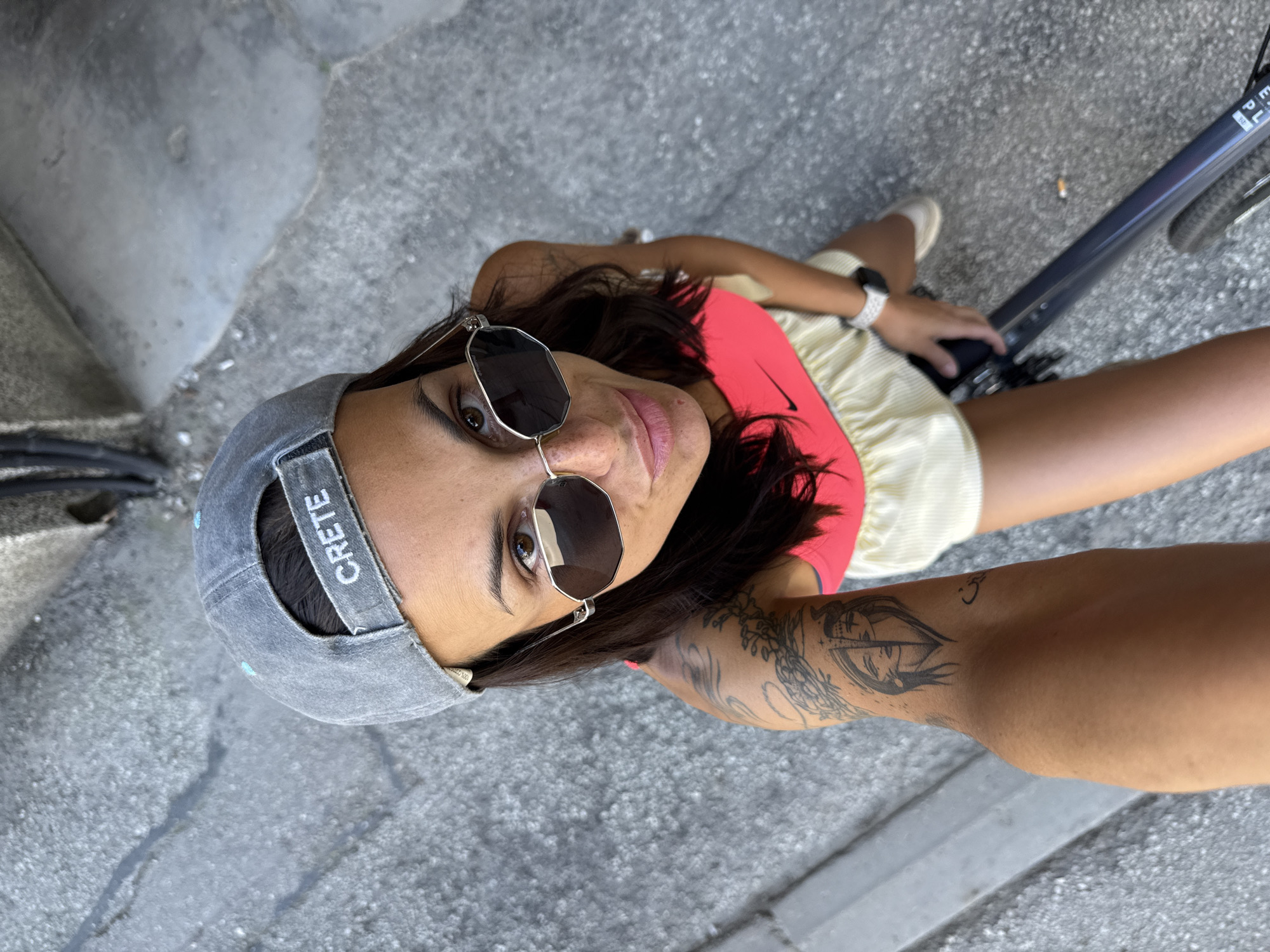 A woman wearing a gray baseball cap that says "CRETE", sunglasses, and a pink sports bra with a yellow skirt is taking a selfie while sitting on a bicycle on a concrete sidewalk. Her right arm, with a tattoo of a face, is extended to capture the photo. The background shows a gray concrete surface and what appears to be a building in the upper portion of the image. The location is the city of Pleven, Bulgaria.