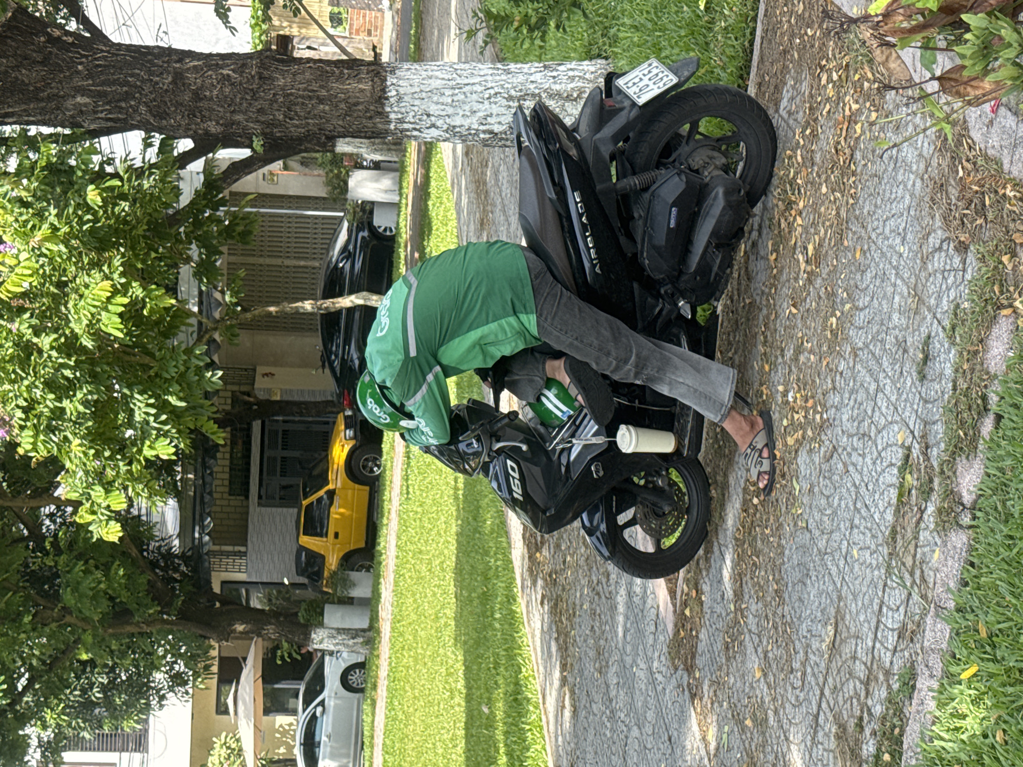 A delivery driver wearing a green Grab uniform is servicing a black motorcycle on a paved sidewalk in a residential area of Đà Nẵng, Vietnam. The motorcycle has a license plate reading "75669 13-92". The driver is bent over the front of the motorcycle, appearing to work on it. There is a cylindrical white object strapped to the front of the motorcycle. In the background, there are trees, a lawn, houses, and parked vehicles, including a yellow car. The sidewalk is gray and patterned, with scattered leaves.