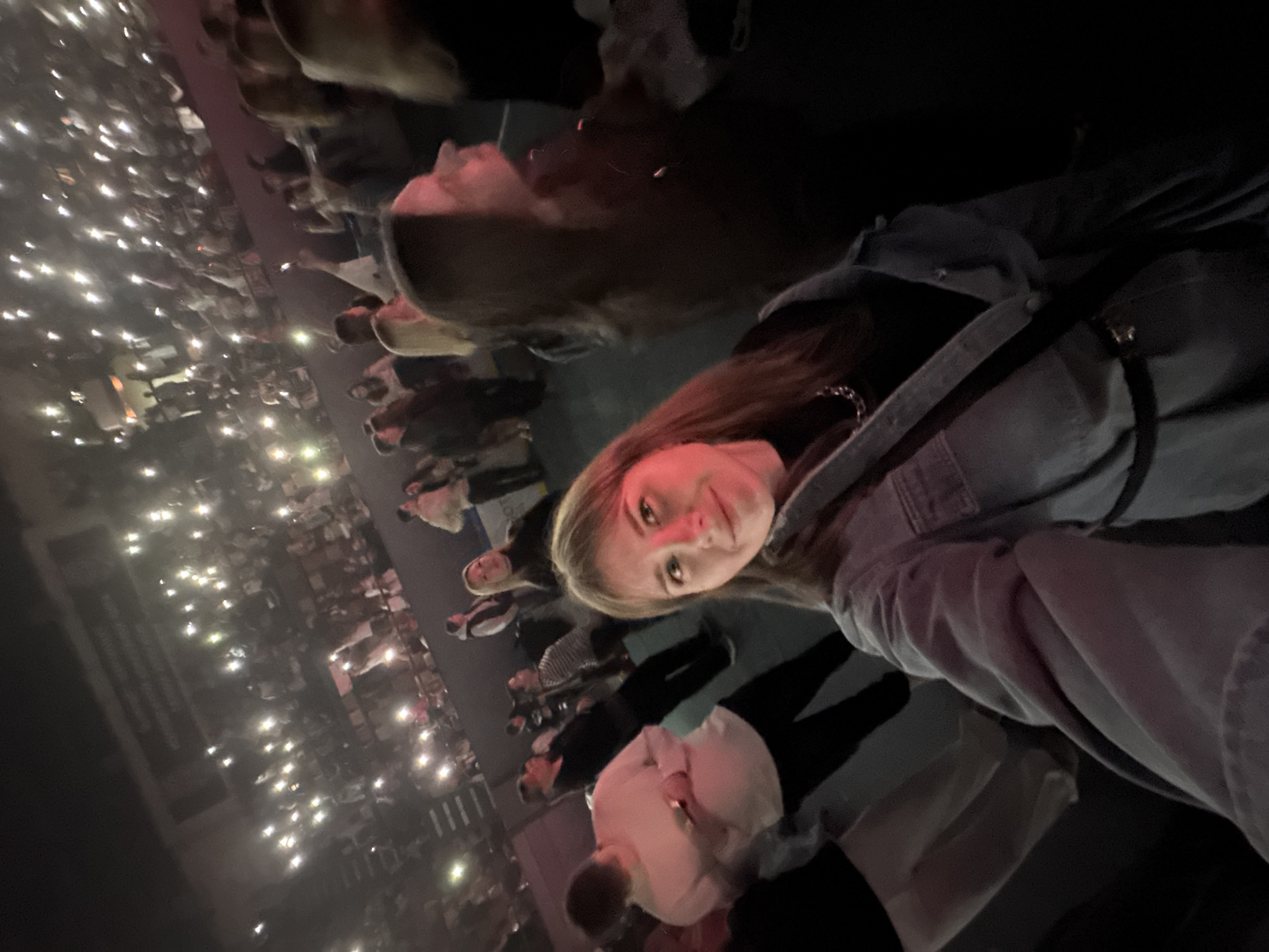 A young woman with long brown hair, wearing a denim jacket, is taking a selfie. She is at a concert, as indicated by the crowd behind her holding up cell phone lights. The environment is dark, but the lights illuminate the crowd and suggest a large venue. The location is identified as Perm, Russia.