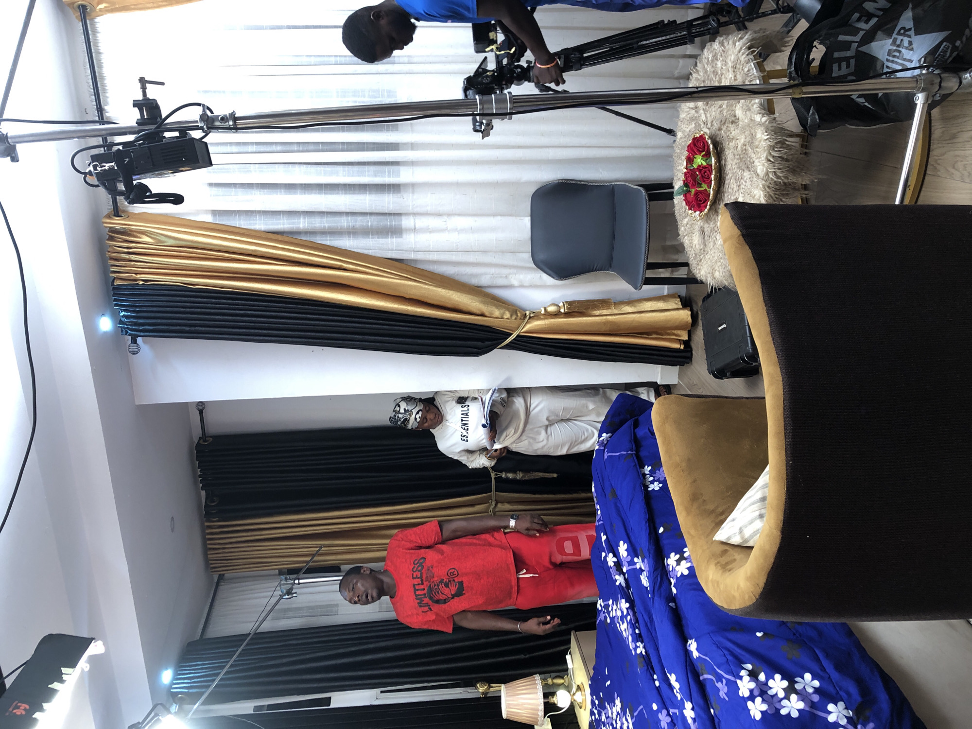 The image captures an indoor scene set up for a professional video or photo shoot, featuring individuals and an array of filmmaking equipment. The room appears to be a residential or studio space.
In the lower portion of the frame, a person wearing a red t-shirt with "LIMITLESS" printed on it and matching red pants stands prominently, looking towards the left. Behind them, another individual in a white hoodie, with the brand "ESSENTIALS" visible, holds a sheet of paper or a script. Adjacent to these people, a bed is made with vivid blue bedding patterned with white and black flowers. A brown armchair is partially visible in the bottom right corner, and a bedside lamp with a pleated shade sits on a nearby table.
The upper and central parts of the image are dominated by the production setup. A person in a blue shirt operates a camera mounted on a tripod or dolly, positioned near a window covered by white blinds. A long metal pole, likely a boom arm or support for lighting, extends across the room, secured by clamps and bearing a small black light fixture. Another larger softbox light is visible in the bottom left, illuminating the scene.
The room's decor includes floor-length curtains with alternating black and gold fabric panels, tied back with a decorative gold tassel. A modern gray chair and a small, shaggy light-colored rug with a golden bowl of red decorative roses on it are also part of the set. Various black equipment cases and bags, one with "IMPER" (possibly "JUMPER" or "IMPERIAL") visible, are scattered on the floor, indicating an active production environment. The lighting suggests artificial illumination is heavily in use.