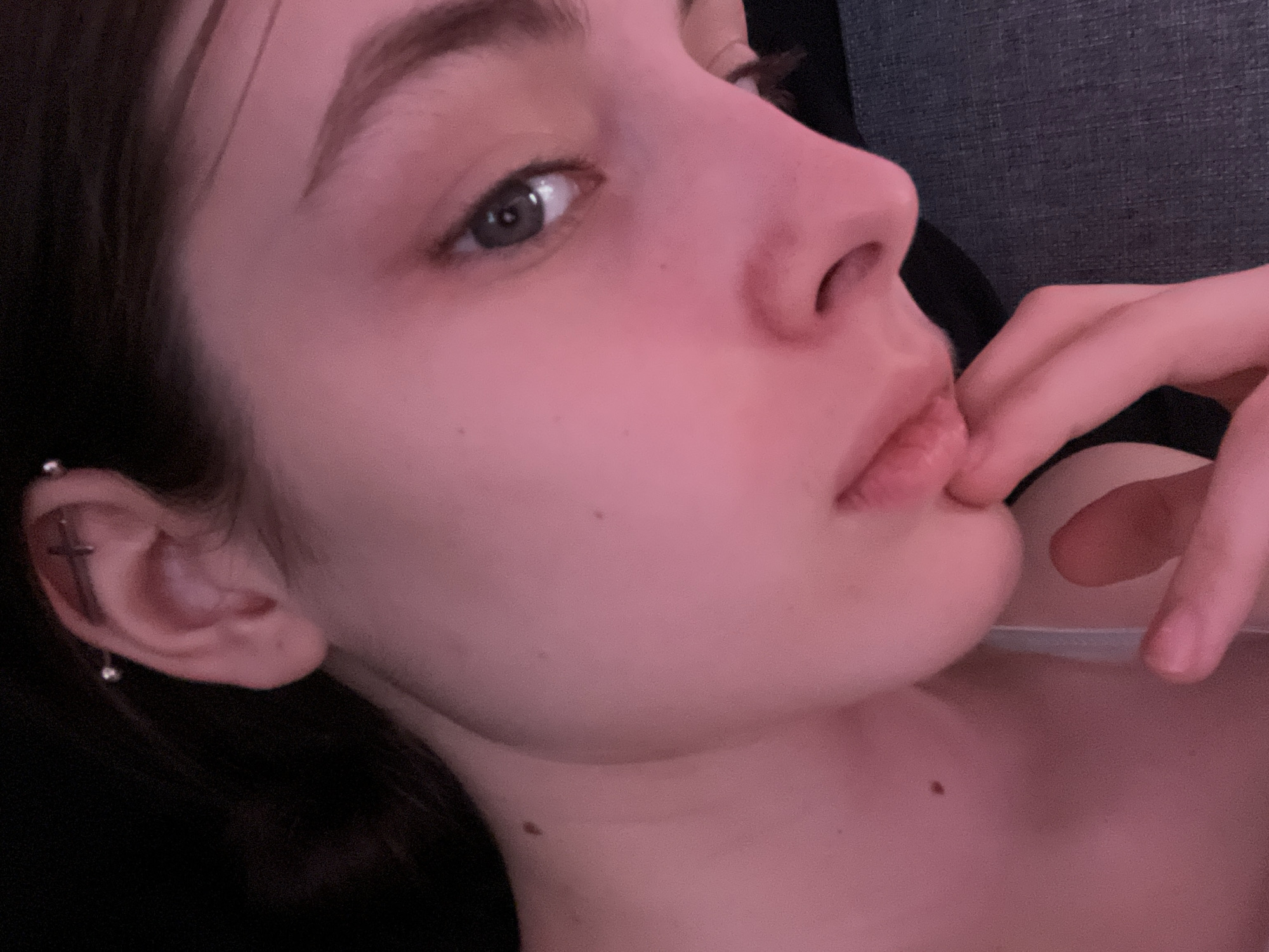 A close-up portrait of a young woman with fair skin, light-colored eyes, and dark hair is featured. She is lying down, with her head tilted back. Her right hand is raised to her mouth, with her index finger touching her lower lip. The ear has two piercings in addition to a cartilage piercing. A mole is visible on her neck and the side of her face. The background is blurry, suggesting she is indoors on a couch or bed. There is no indication of this picture being taken in St. Petersburg, Russia.