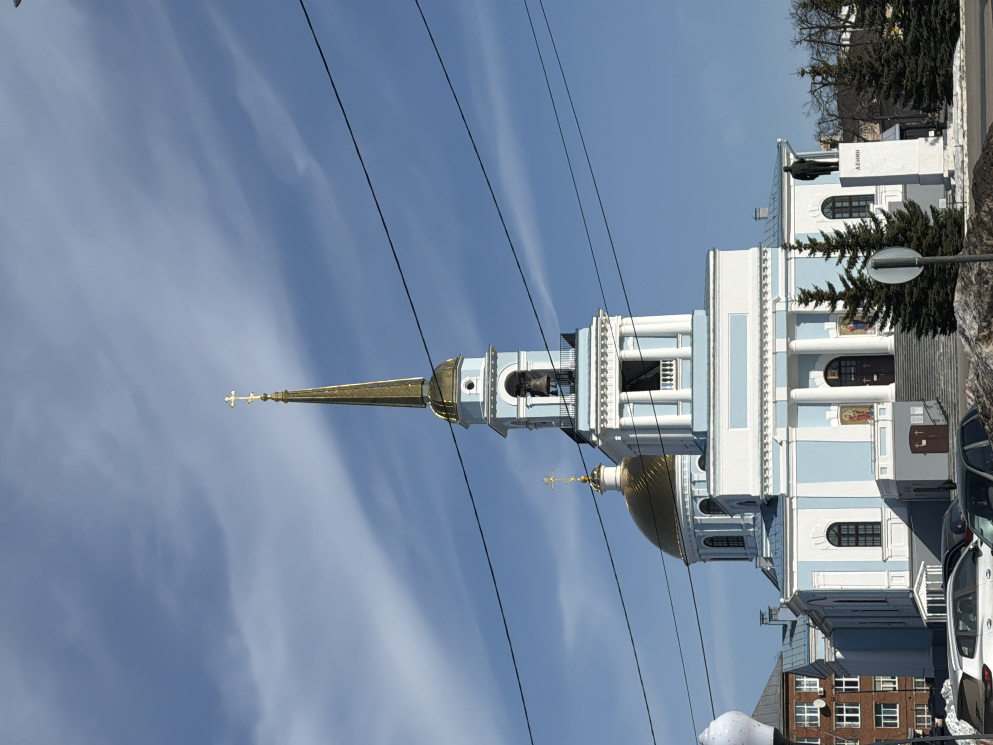 The image shows the Ascension Cathedral in Votkinsk, Russia. The cathedral is a light blue and white building with a prominent gold dome and a tall spire topped with a cross. Power lines are visible in the foreground, crossing the sky. A statue of Lenin is seen to the right of the building. Several trees and cars are also visible. The sky is a clear blue with wispy clouds.