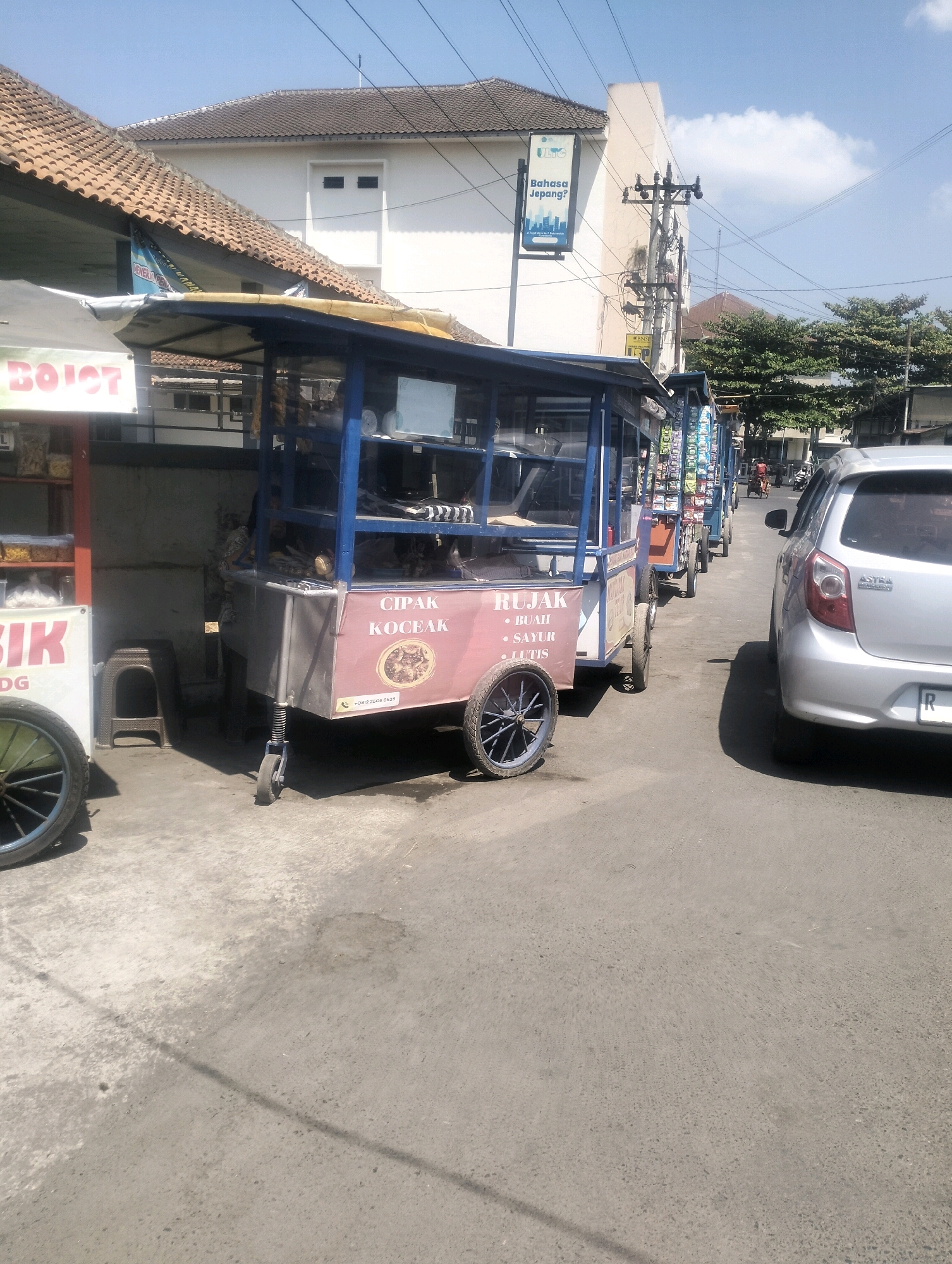 The image captures a street scene in Karangsoka, Indonesia, on a bright, sunny day. The foreground is dominated by a row of food carts, the closest one being pink with blue trim and a sign listing "CIPAK, KOCEK, BUAH, SAYUR, LUTIS". This cart also has a phone number printed on it: "+0812 2506 6525". A silver sedan is parked on the right side of the street, facing away from the camera. Behind the food carts, a white building with a tiled roof is visible, featuring a blue and white sign that says "Bahasa Jepang?". A utility pole with numerous wires stands tall against the clear blue sky. The ground is paved with asphalt, showing some wear and tear. Several trees with lush green foliage line the street in the background. The overall impression is one of a typical day in a lively, urban environment.