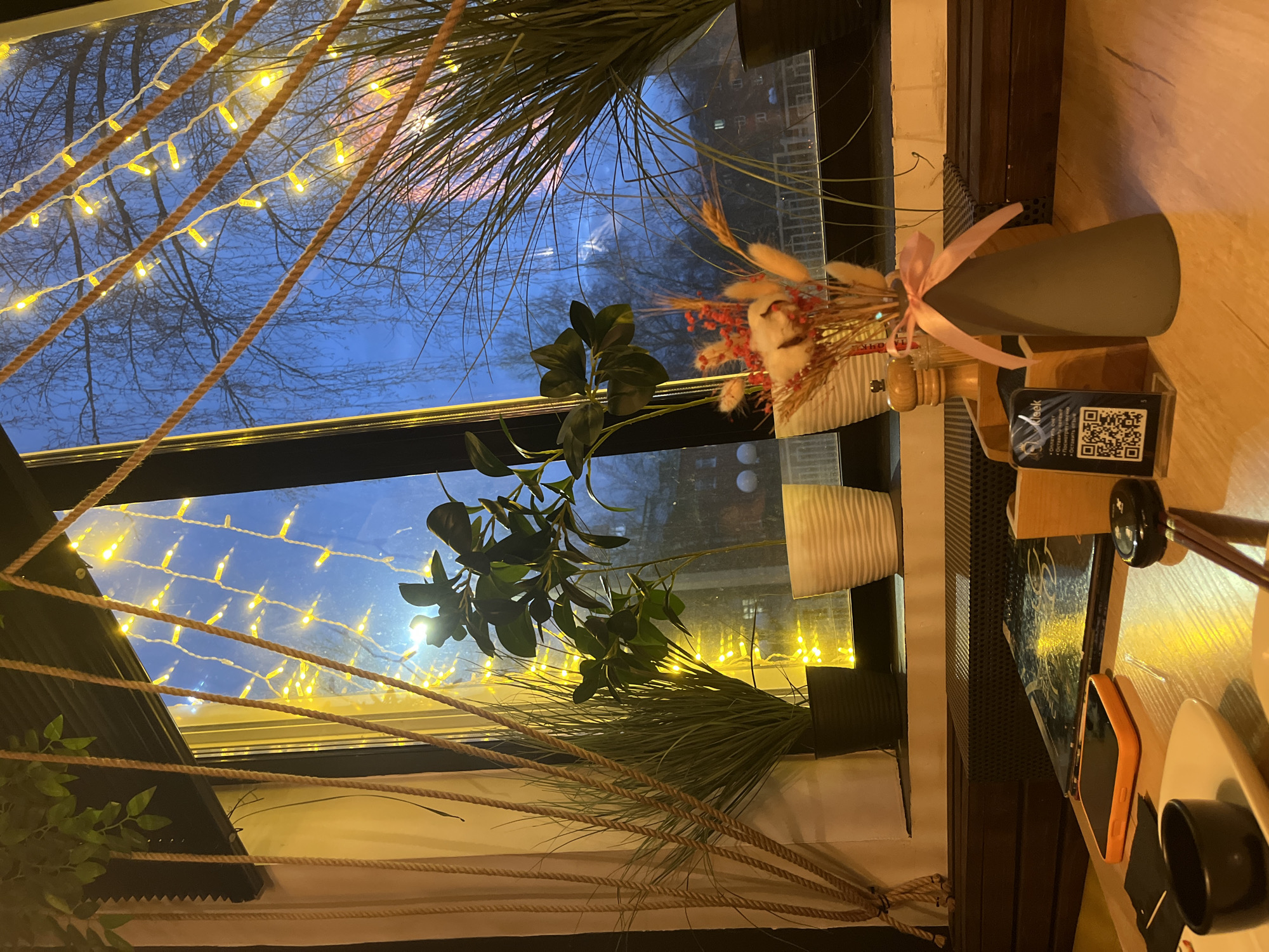 The image depicts a cozy interior scene viewed through a window during the evening or night. Warm yellow fairy lights are strung horizontally across the window pane, interspersed with thick, braided ropes, casting a soft glow. Beyond the window, the sky is a deep blue, and the silhouetted branches of bare trees are visible, suggesting an autumn or winter season.
Inside, on what appears to be a wooden table or wide windowsill, a collection of objects creates a natural and inviting atmosphere. A large green leafy plant sits prominently near the window, alongside strands of long, grass-like green foliage. Further to the right, a dried floral arrangement, containing red berries, fluffy white elements (possibly cotton or dried flowers), and dried grasses, is nestled in a small pot and adorned with a pink ribbon. Behind this, a dark grey or green conical vase, also tied with a pink ribbon, adds to the decor.
On the wooden surface, a QR code is displayed on a small stand. Above the code, the word "kreek" is visible. Below the QR code, text in Russian begins with "Осеннее...", repeated multiple times, likely indicating an "Autumn menu" or seasonal offering relevant to the location context of Izhevsk, Russia. Adjacent to these items, a smartphone in an orange case rests on the table, next to a dark-colored cup on a white saucer. Other details include a wooden pepper mill, a black speaker grille, and what appears to be a book or tablet. The overall setting evokes a comfortable and relaxed ambiance, characteristic of a cafe or restaurant.